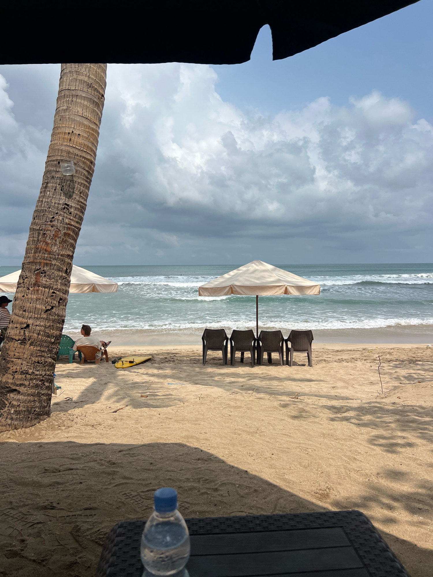 A serene beach scene unfolds in Kuta, Indonesia, under a cloudy sky. A tall palm tree stands to the left, casting a shadow. In the background, the ocean waves gently roll onto the sandy shore, with two beach umbrellas providing shade. Four empty chairs are arranged neatly in front of one of the umbrellas. A person sits in a chair near the water, engrossed in a book, with a yellow surfboard beside them. In the foreground, a bottle of water rests on a dark surface.