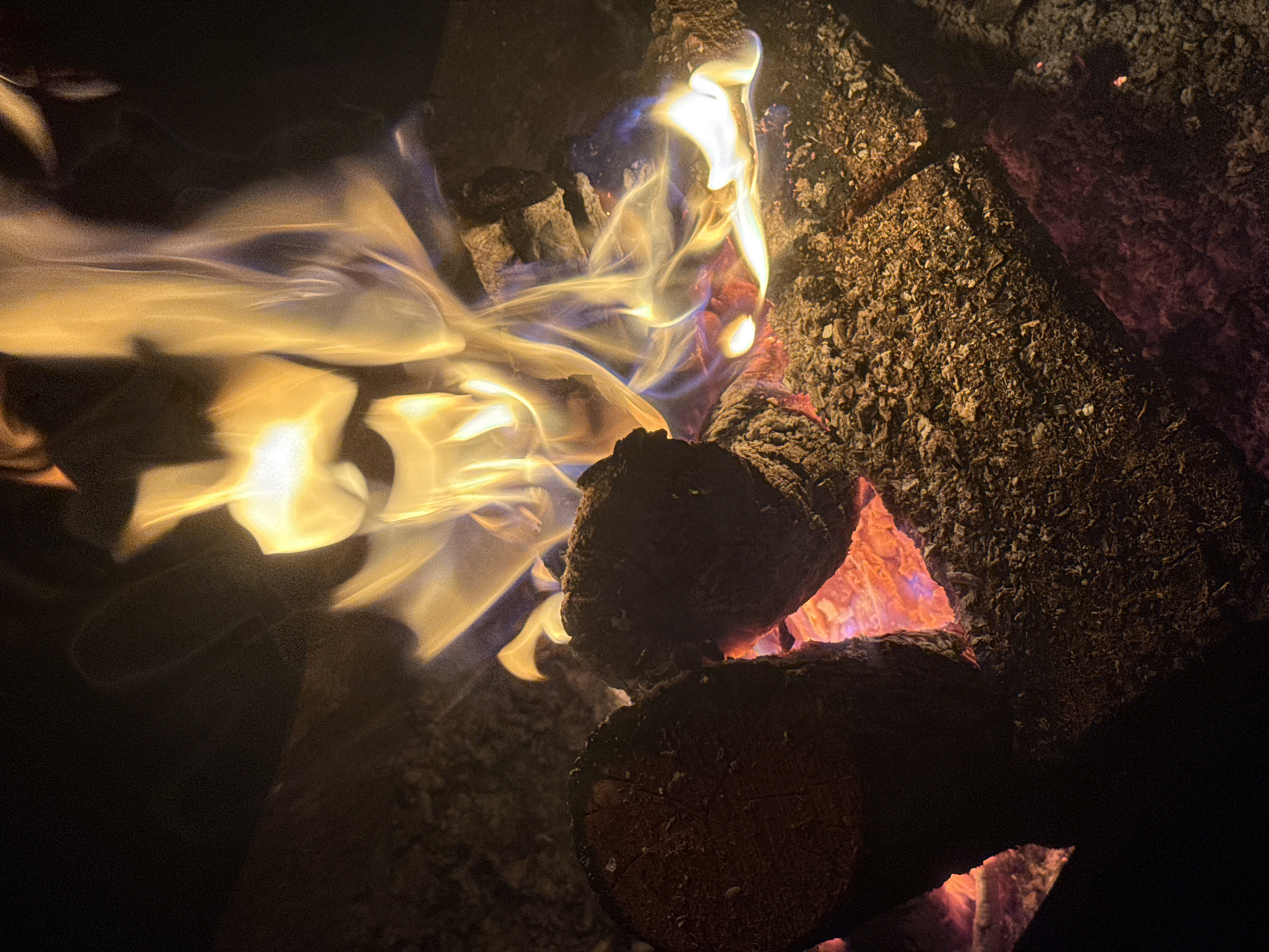 The image depicts a close-up, low-light view of a bonfire in Автомобилист-1 Снт, Russia. Two logs are prominently positioned in the foreground, with a glowing bed of embers beneath and between them. The logs are dark and appear to be burning. Flames, characterized by vibrant yellows, oranges, and hints of blue, rise dynamically from the wood and embers, illuminating the scene. The background is dark, suggesting nighttime or a shaded environment, with the focus entirely on the fire and its immediate surroundings. No people are visible in the frame. The overall impression is one of heat and light emanating from a controlled fire.