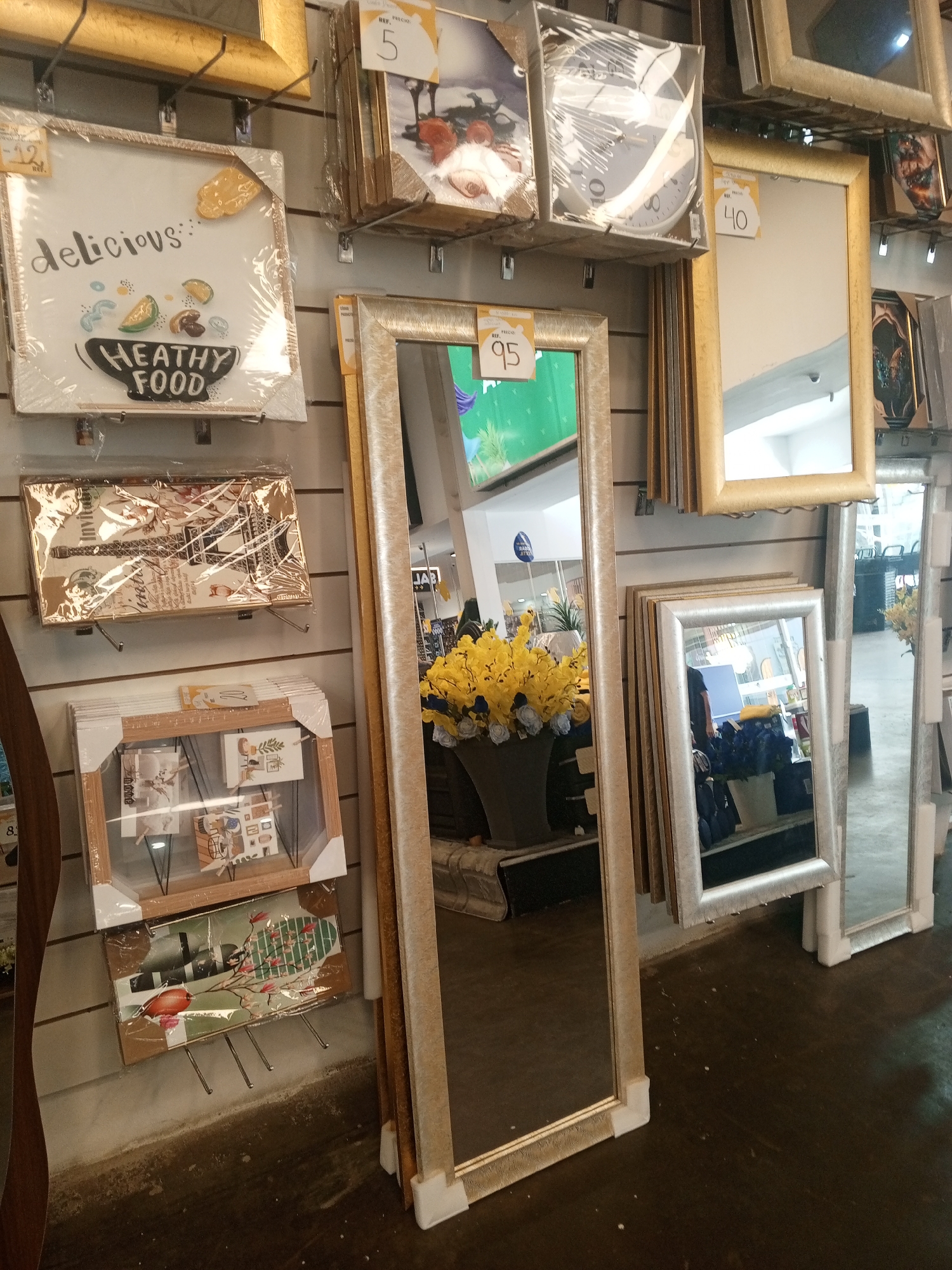The image depicts a retail display of home decor items in what appears to be a store. The scene is likely located in Pampatar, Venezuela.
The display features a variety of framed art, mirrors, and decorative objects hung on a wall. A prominent element is a series of tall, narrow mirrors with metallic gold frames. Some of the art pieces are still encased in plastic wrapping, suggesting they are new inventory. There are framed pictures with food-related themes, Eiffel tower art and other wall decorations.
There are yellow flowers in a vase near the center of the image. The floor is dark and appears to be made of wood or a similar material.