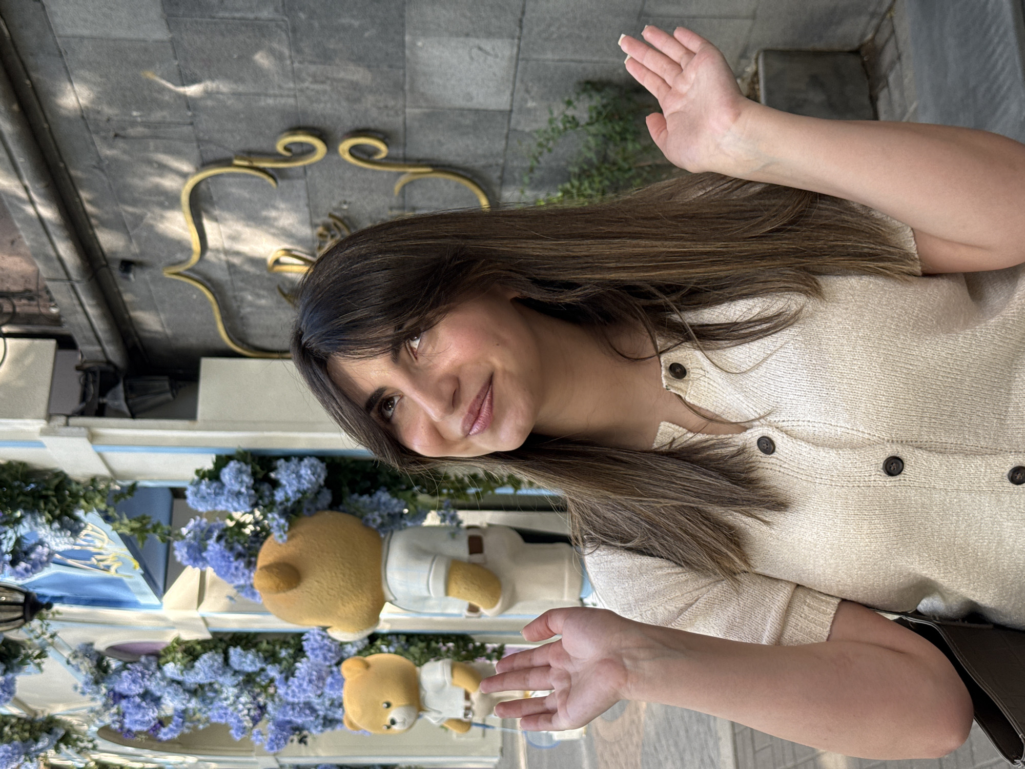 A young woman with long, dark brown hair and a friendly smile poses for the camera in an outdoor setting in Yerevan, Armenia. She is wearing a light-colored, possibly cream or beige, knitted cardigan with dark buttons. Both of her hands are raised in a welcoming or gesturing manner, with palms open.
The background features a grey stone or concrete wall with ornate golden metallic decorative elements. Daytime lighting casts distinct shadows, indicating a sunny day. To the woman's left and below her, two light brown teddy bears, one larger and one smaller, are visible. Both bears are dressed in white shirts and are situated amidst abundant blue flowers, likely hydrangeas, arranged as part of a decorative display. This floral and teddy bear arrangement suggests a charming shop front, cafe entrance, or a specially decorated public space. No visible text is present in the image.
