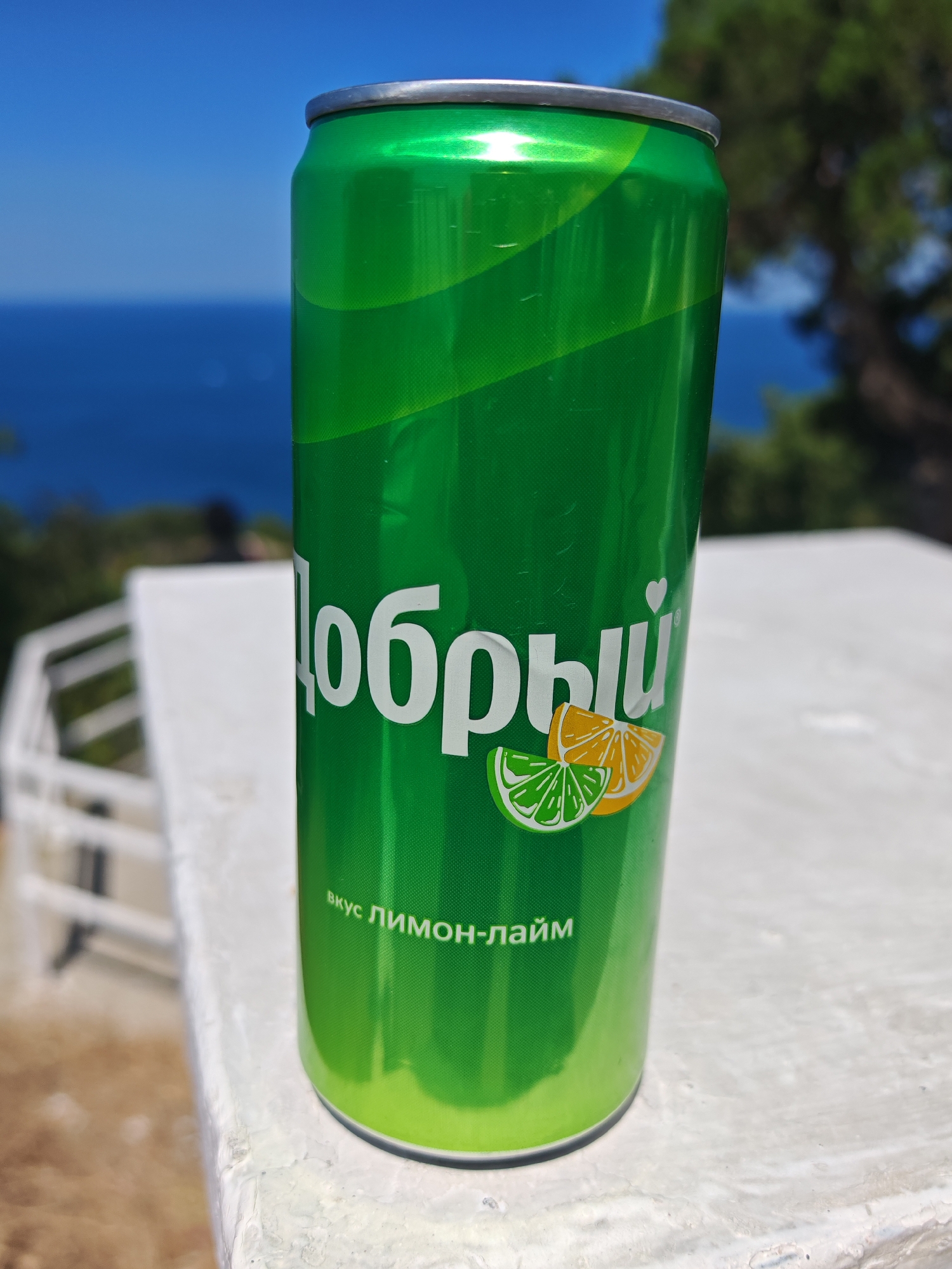 A bright green soda can with white Cyrillic lettering and citrus fruit illustrations sits on a white, textured surface. The can has the word "Добрый" (Dobryi) prominently displayed, along with images of lime and orange slices. Below these, smaller text reads "вкус лимон-лайм" (lemon-lime flavor). The background is blurred, showing a vibrant blue sky meeting the sea, with hints of green foliage and a white structure, suggestive of a scenic outdoor location. The lighting indicates a sunny day. The image appears to be taken in Sukko, Russia, given the context provided.