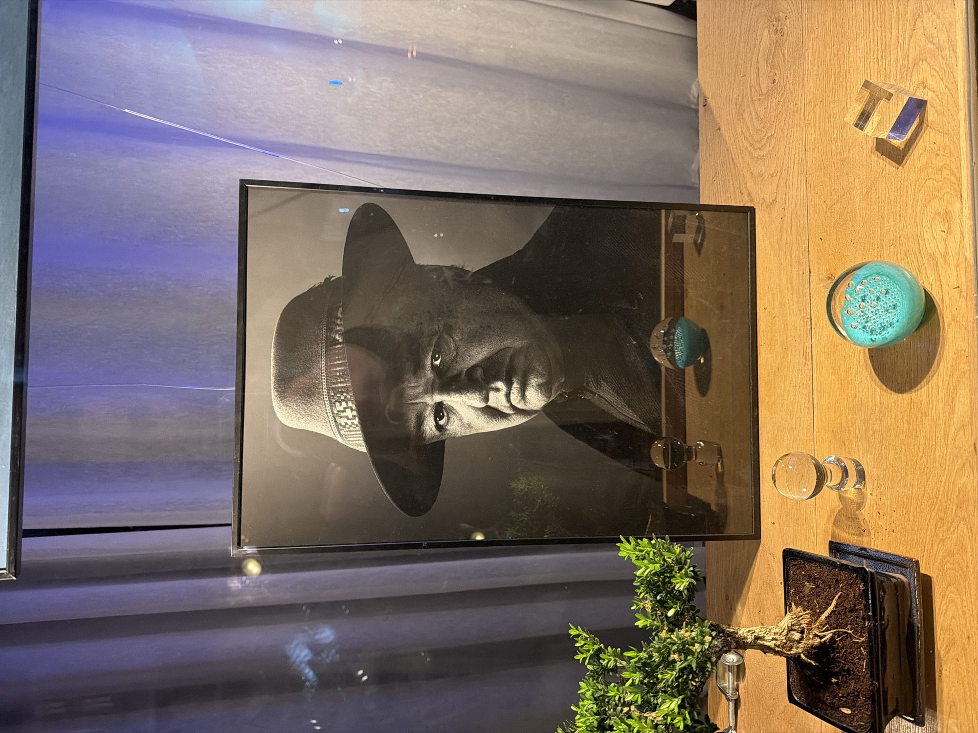 The image depicts a black and white portrait of a man wearing a hat displayed in a room with various decorative objects. The portrait is framed and positioned against a reflective surface, possibly a window or glass panel. To the right of the portrait is a wooden panel featuring decorative items, including a clear cube, a turquoise glass orb, a clear glass doorknob, and a small bonsai tree in a black pot. The background appears to show a slightly blurred cityscape.
There is no definitive evidence to pinpoint the precise location within Paris.