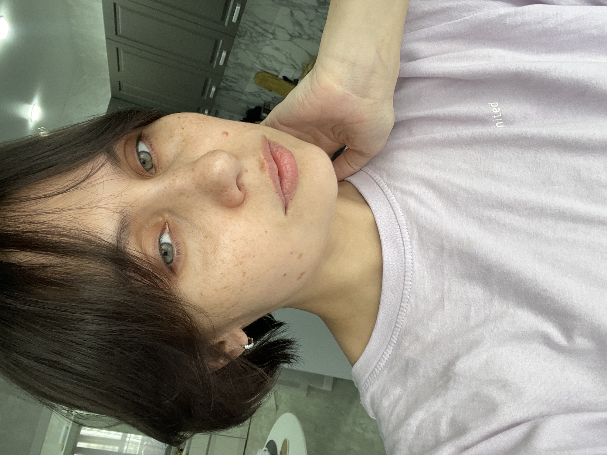 A close-up portrait shows a woman with fair skin and freckles, wearing a light purple t-shirt, posing indoors. The t-shirt has the word "nited" printed on it. She has short, dark brown hair and light green eyes. Her right hand is positioned near her face, touching her neck. A silver hoop earring is visible in her left ear. The background appears to be a room with furniture and a window. There is no specific evidence in the image to indicate that it was taken in Izhevsk, Russia.