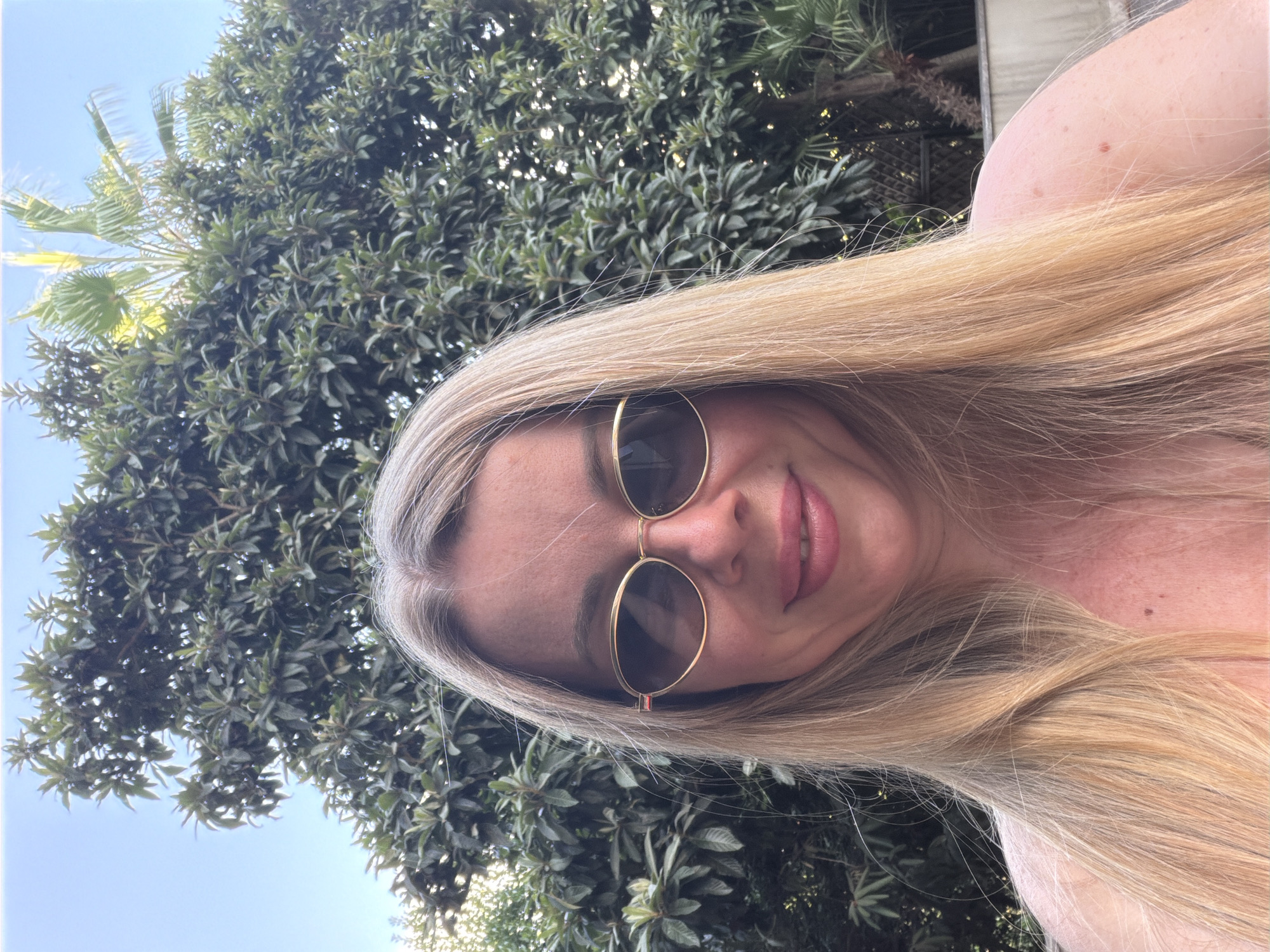 A blonde woman is posing for a selfie outdoors. She has long blonde hair, is wearing sunglasses, and has pink lipstick on. She is smiling and looking at the camera. The background is a lush green tree with a clear blue sky above. There is a palm tree visible in the upper right corner of the image. The lighting suggests it is daytime. No other people are visible in the image. The location is not discernible from the image.