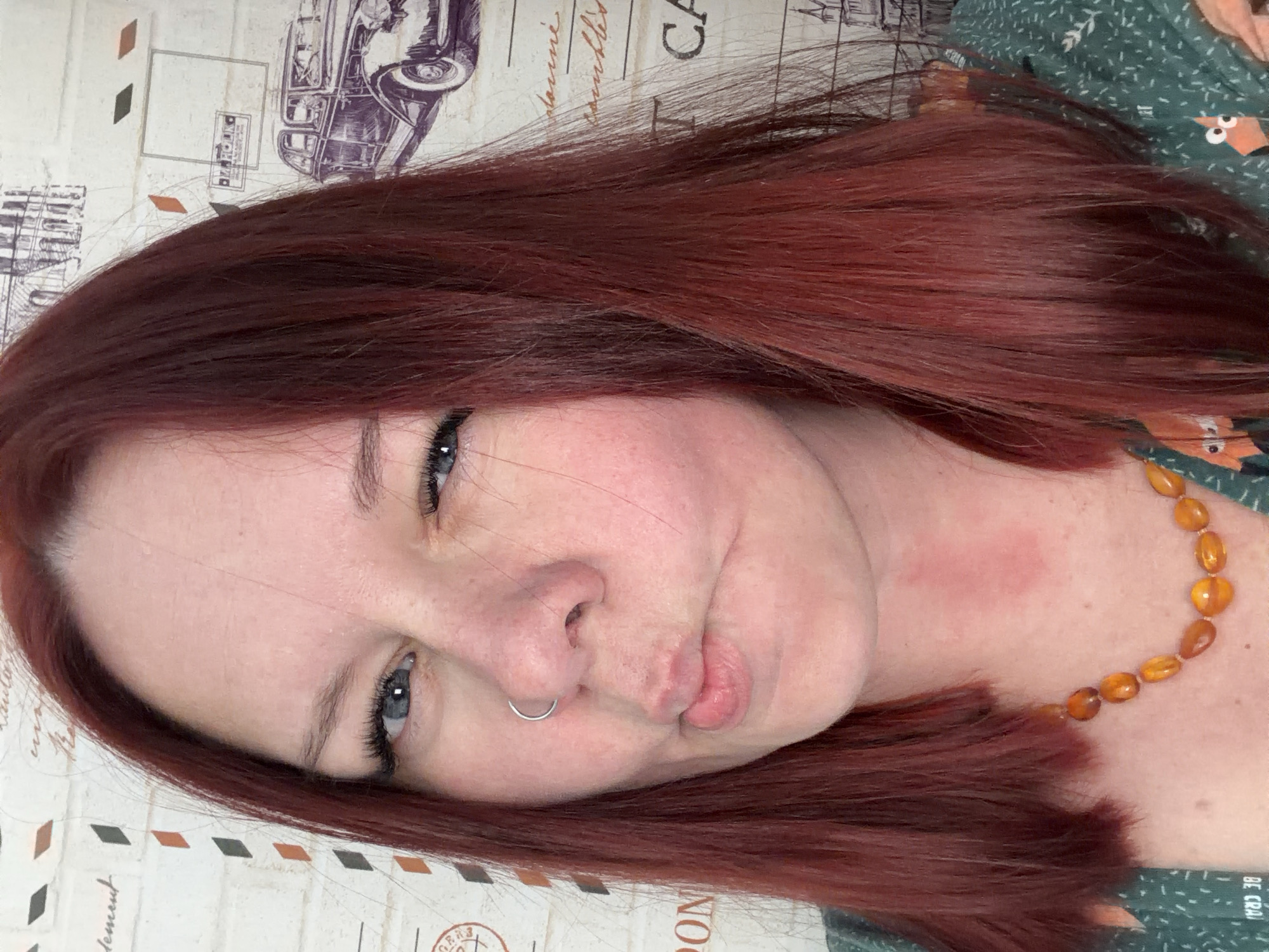A woman with auburn hair is shown in a close-up selfie. She has fair skin, blue eyes, and a silver nose ring. She is making a pouting face. A reddish mark is visible on her neck. She is wearing a necklace of amber-colored beads. She is wearing a green shirt with a pattern of orange foxes. The background is a beige surface with a pattern that includes an old car illustration and script. There is no indication of the city of Chaikovsky, Russia in the image.