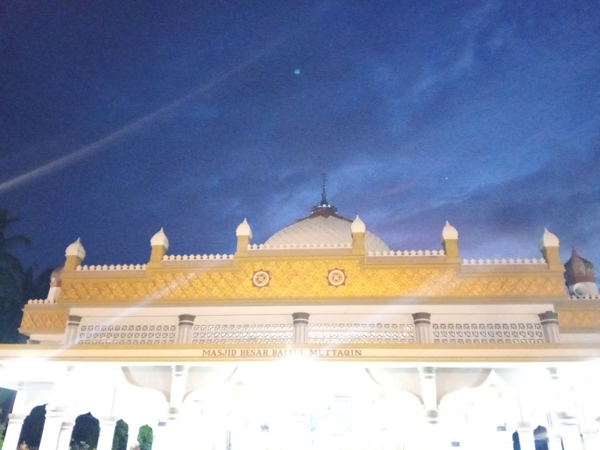 The image captures the facade of Masjid Besar Baitul Muttaqin, a large mosque located in Uteunghatom, Indonesia, at dusk. The sky is a deep, darkening blue, with wisps of clouds and a faint contrail or light streak visible. The mosque's architecture is prominent, featuring a golden decorative frieze with intricate patterns, topped with small white finials. Below this, a white architectural element with repeating decorative motifs and dark columns stands out. A prominent inscription in red lettering reads "MASJID BESAR BAITUL MUTTAQIN". The foreground shows the illuminated entrance area of the mosque, with arched openings and bright white lighting, suggesting it is open and in use. The overall scene conveys a sense of evening tranquility and the presence of a significant religious building within its urban context, although no people are clearly visible in the frame.