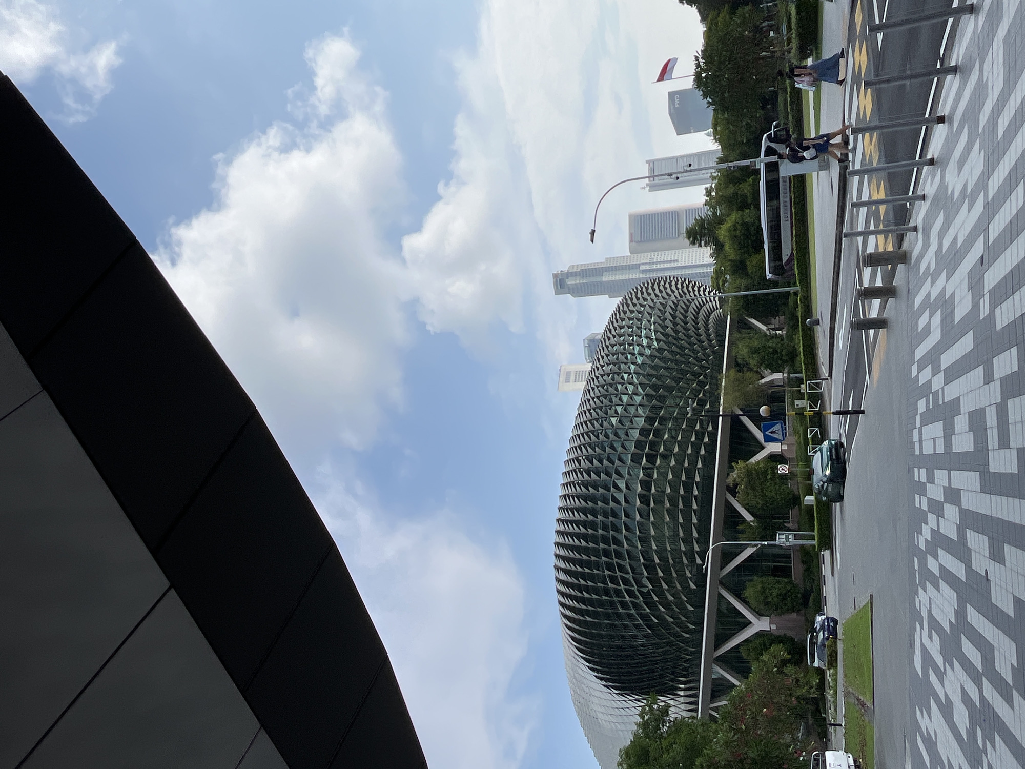 The image depicts a street scene in Singapore, with the iconic Esplanade - Theatres on the Bay in the background. The building, known for its distinctive durian-like architectural design, is a prominent feature of the Singapore skyline. In the foreground, a modern pedestrian crossing with a patterned pavement is visible. Two individuals are crossing the street from right to left, and another person is walking in the same direction further to the right. A white luxury coach bus is parked on the left side of the road. Several cars are also present on the road, and a white pickup truck is parked on the far left. The sky is partly cloudy, with a clear blue expanse in between. Tall buildings, including skyscrapers, rise in the distance, indicative of a bustling urban environment. A flag, possibly the Singaporean flag, is visible on a pole in the distance. The scene suggests a bright, possibly late afternoon, day in Singapore.