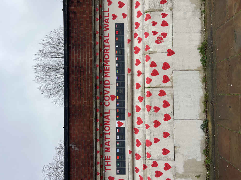 The image displays a section of The National Covid Memorial Wall, located along the Embankment in London, United Kingdom. The wall consists of two primary architectural sections: an upper layer of red brick, and a lower, prominent section constructed from light-colored, possibly cream or pale grey, stone blocks.
Above the stone section, "THE NATIONAL COVID MEMORIAL WALL" is inscribed prominently in red capital letters. Directly beneath this text, a horizontal row of eighteen black rectangular plaques is affixed to the wall. Each plaque displays a national flag, with the United Kingdom flag visible at both ends of the row and other international flags spanning between them. A single, larger red heart plaque is centrally positioned within this row, replacing one of the national flag plaques.
The substantial lower section of the light-colored stone wall is extensively adorned with hundreds of individual red paper or card hearts of various sizes. These hearts are affixed to the stone surface, and many exhibit visible handwritten messages or names. The hearts are distributed unevenly, with clusters and sparser areas.
At the base of the wall, a paved walkway, composed of large, rectangular stone slabs, is visible. Sparse green vegetation, consisting of small plants and weeds, grows in the crevices where the wall meets the pavement. Above the red brick section, the bare branches of deciduous trees are visible against a uniformly overcast, light grey sky. No individuals are present in the scene.