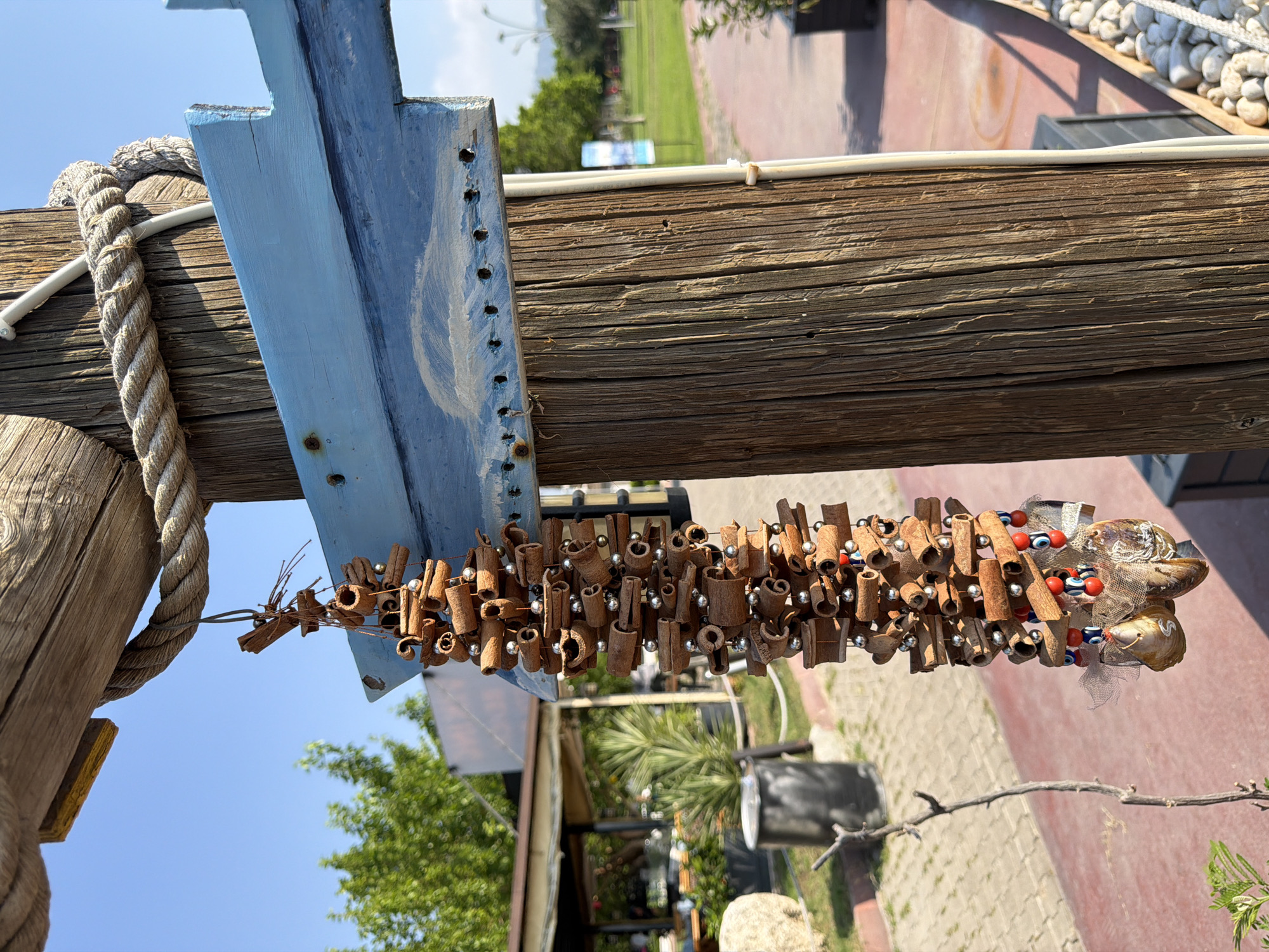 The image shows a close-up of a rustic wooden post in Kemer, Turkey, with a hanging decoration made of cinnamon sticks and beads. The decoration is suspended from a blue wooden plank attached to the post, which is itself wrapped with a thick rope. In the background, a paved walkway with a red surface is visible, leading towards a garden area with trees and outdoor seating. The sky is clear and blue. The overall scene suggests an outdoor setting, possibly a restaurant or cafe, with a focus on natural materials and a laid-back atmosphere.