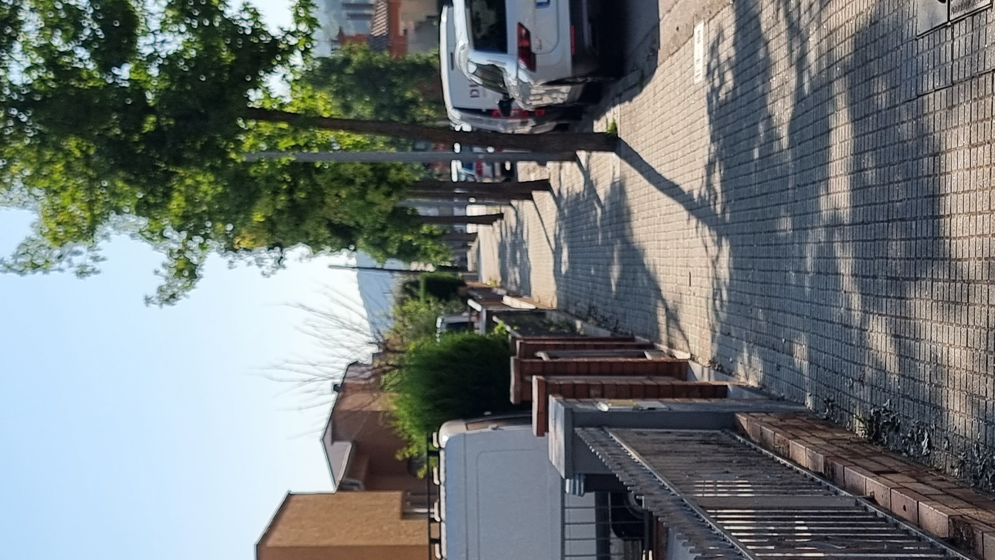 The image shows a sun-drenched street in Sant Joan Les Fonts, Spain, with a paved sidewalk lined by trees and parked vehicles. The sidewalk, made of square tiles, is shaded by the canopy of leafy trees, casting dappled shadows across its surface. On the left side of the sidewalk, there is a metal fence with a brick base, separating it from a property with a white van. Beyond the van, a brick wall with brick pillars runs along the sidewalk. Further down the street, more parked cars are visible, including a white SUV on the right. Buildings with reddish-brown roofs are present on the left and right sides of the street, receding into the distance. The sky is clear and blue, indicating fair weather.
