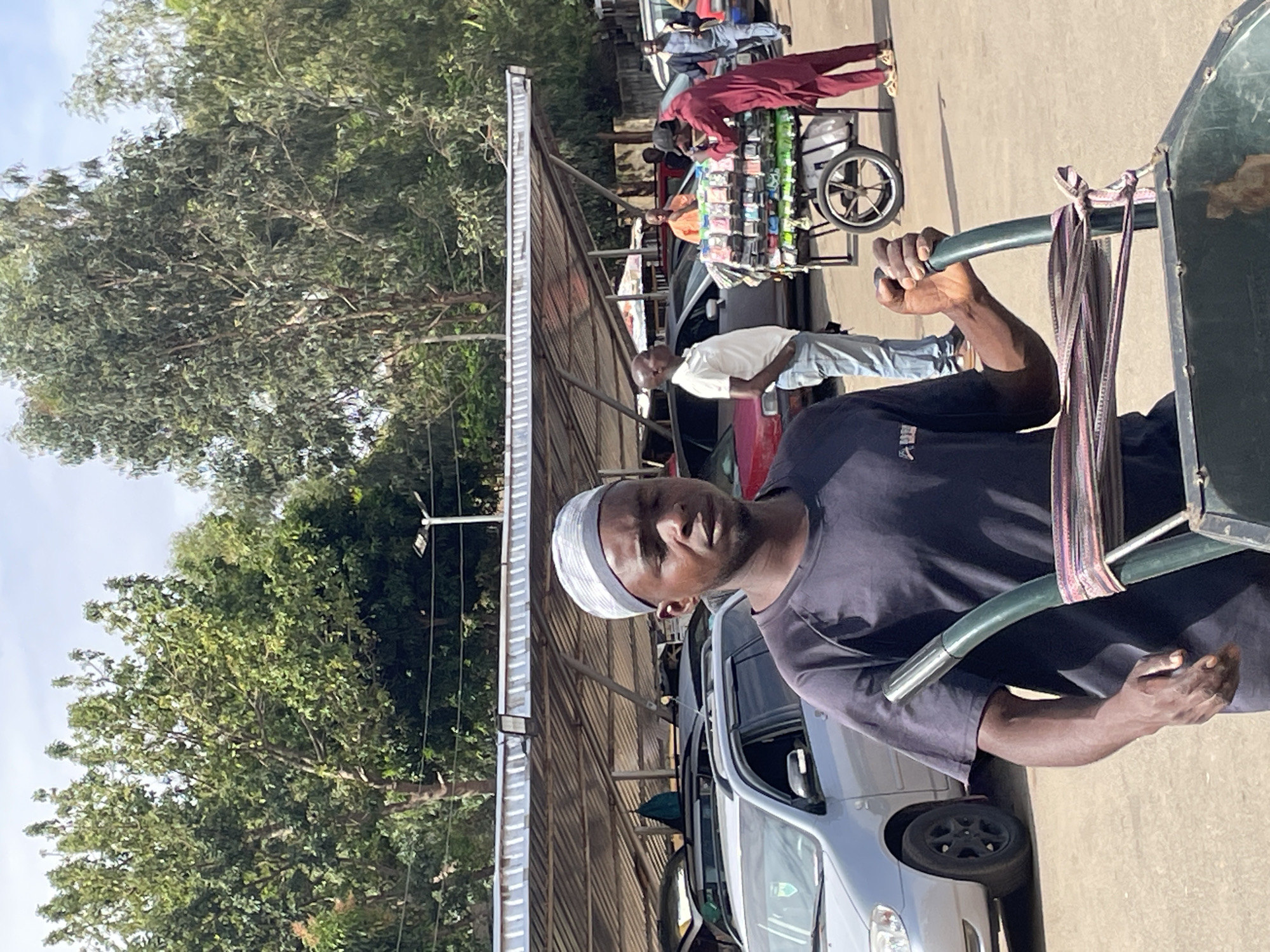 The image shows a man standing in Bukuru, Nigeria. He is wearing a dark t-shirt and a white cap. He is holding the handles of a green pushcart, with striped fabric tied to it. In the background, there are other people, vehicles, and a vendor's cart selling small items. Trees and a corrugated metal roof structure are also visible. The scene appears to be a street or market area.