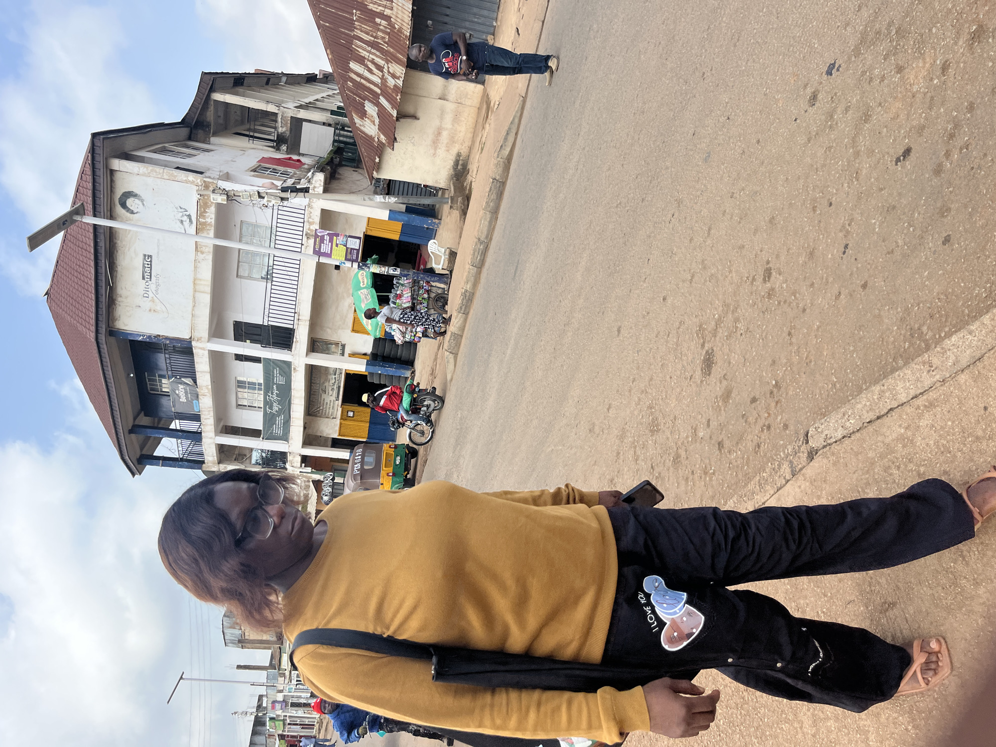A woman wearing a yellow long-sleeved shirt and black pants with a heart design is walking on a dirt road in the city of Gura Top, Nigeria. She is looking down and holding a phone in her right hand. In the background, there is a leaning building with a sign that reads "Ditomatic" and "Photography." Several vehicles, including a tricycle and motorcycles, are present on the road, along with other people. A man in a dark t-shirt and jeans is standing on the side of the road, holding a white object. The environment appears to be a busy street with shops and market stalls. The sky is cloudy.