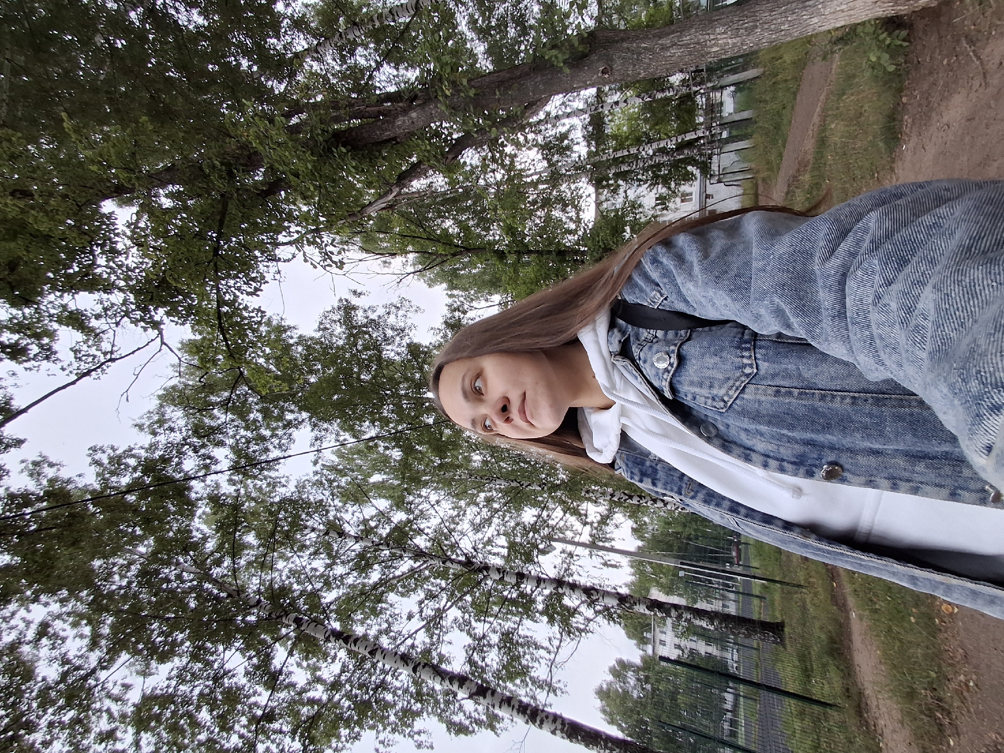 A young woman with long brown hair is standing outdoors, looking upwards. She is wearing a denim jacket over a white hooded sweatshirt. The setting appears to be a park or residential area with numerous trees, including birch trees with distinctive white and black bark. In the background, a fence and a multi-story building are visible, suggesting an urban or suburban environment. The ground is covered with dirt and grass, and some fallen leaves are present. The sky is overcast. This scene is likely located in Chaikovsky, Russia, given the visual cues of the architecture and vegetation.