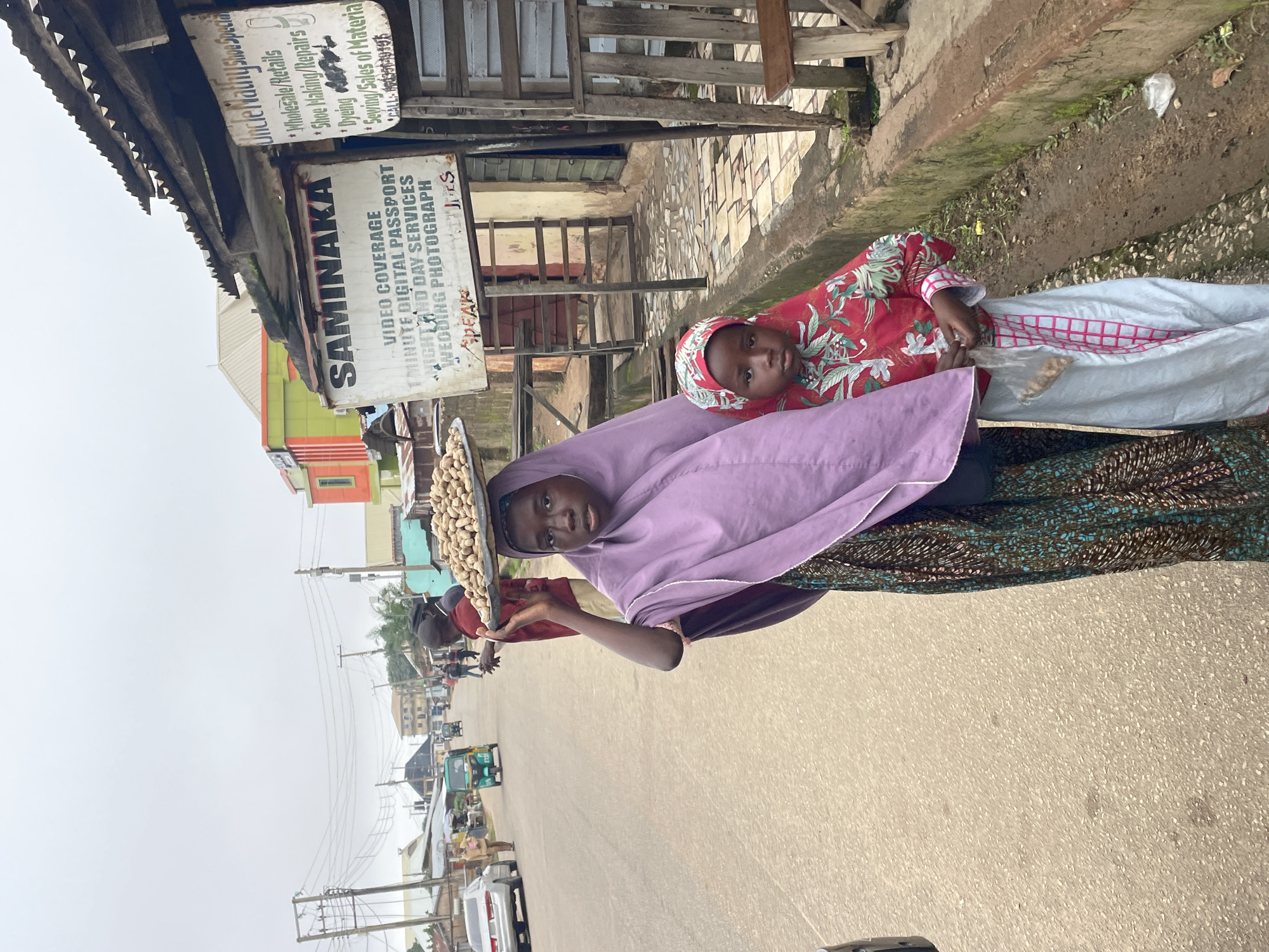 In Jos, Nigeria, on a cloudy day, a young Black girl, likely pre-teen, walks along a paved street with a tray of peanuts balanced on her head. She is wearing a purple hijab and a patterned skirt. Beside her, a younger Black child, possibly a girl, stands dressed in a red and white floral shirt and a light blue and pink checkered skirt and head covering. The younger child holds a clear plastic bag containing peanuts. They are on the right side of the road, with a drainage ditch running alongside the pavement.
Behind them, to the right, is a weathered structure with a corrugated metal roof, displaying several signs. One sign, partially obscured, reads "Uncle Mathy Shoe Specia..." and lists services such as "Wholesale/Retails", "Shoe Making/Repairs", "Dyeing", and "Sewing/Sales of Material". Another larger sign, also weathered, says "SAMINAKA" and advertises "VIDEO COVERAGE", "1 MINUTE DIGITAL PASSPORT", "NIGHT AND DAY SERVICES", and "WEDDING PHOTOGRAPH". A wooden structure, possibly a bench or stall, is visible in front of the building.
Further down the street, other people can be seen walking, and several vehicles are present, including a white car on the left and a green tricycle-like vehicle in the middle distance. The overall environment appears to be an urban setting with buildings lining the street. The sky is overcast, suggesting a generally diffused light.