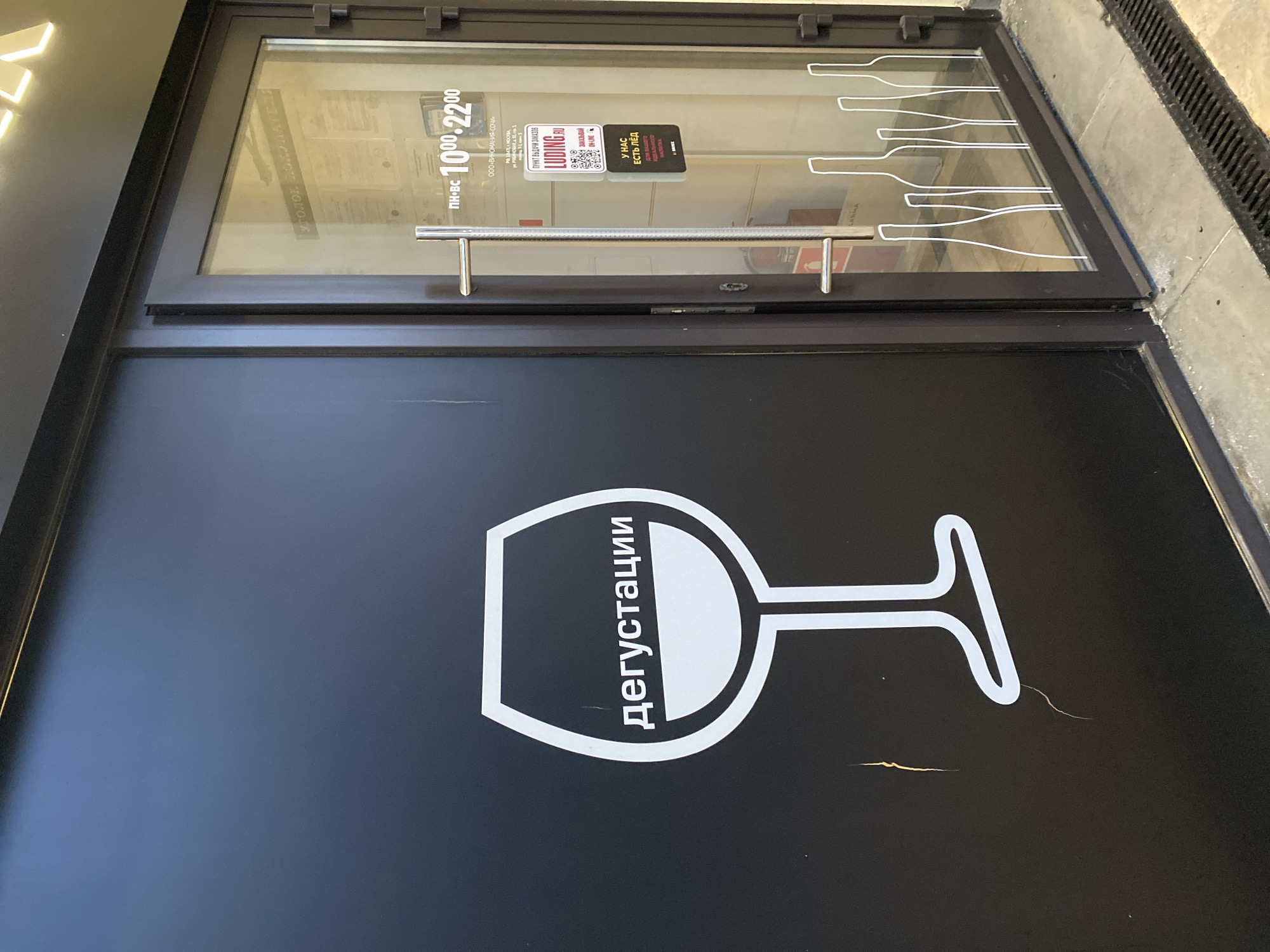 Here is a description of the image:
The image shows a close-up of the entrance to a wine tasting (дегустации) location in Sochi, Russia. The entrance features a black door with a white graphic of a wine glass and the word "дегустации" (tasting) written vertically next to the glass. Above the door is a glass window with various stickers and information posted. A silver horizontal door handle is fixed to the glass door. The stickers include operating hours (10:00-22:00 from Monday to Sunday), information about the company "Vinomanija-Sochi", a QR code linked to "Luding.ru", and a sign that says "We have ice for your ideal drink." There are also white silhouettes of wine bottles decorating the glass. The entrance is located in an urban area with a concrete sidewalk visible on the right side.