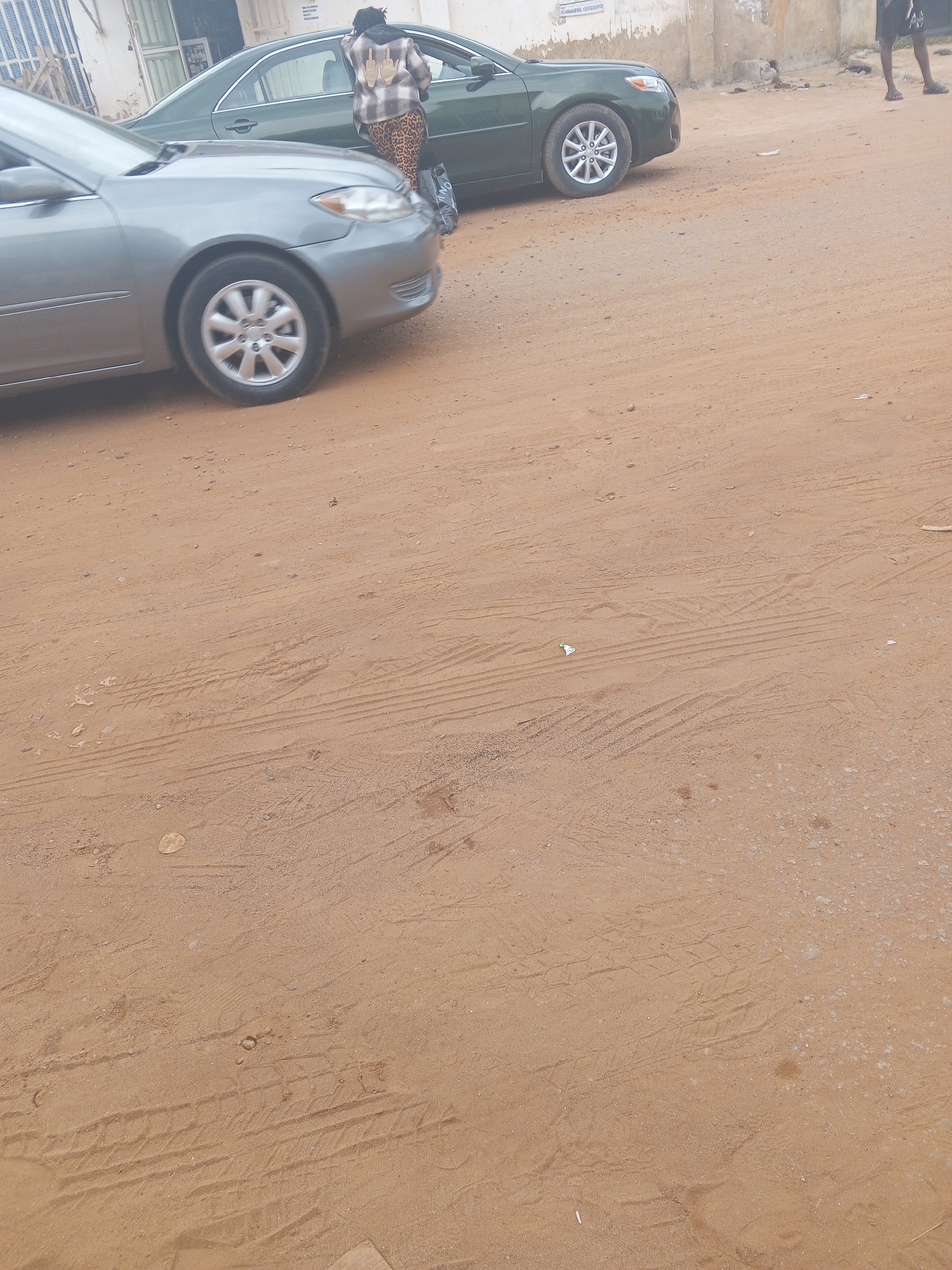 In Abuja, Nigeria, on a dirt road, a person is standing next to a dark green sedan. The person is wearing a patterned jacket and leopard-print pants. A silver sedan is parked in front of the green car, partially obscuring it. The ground is covered in sand and tire tracks are visible. In the background, there is a building with a faded facade.