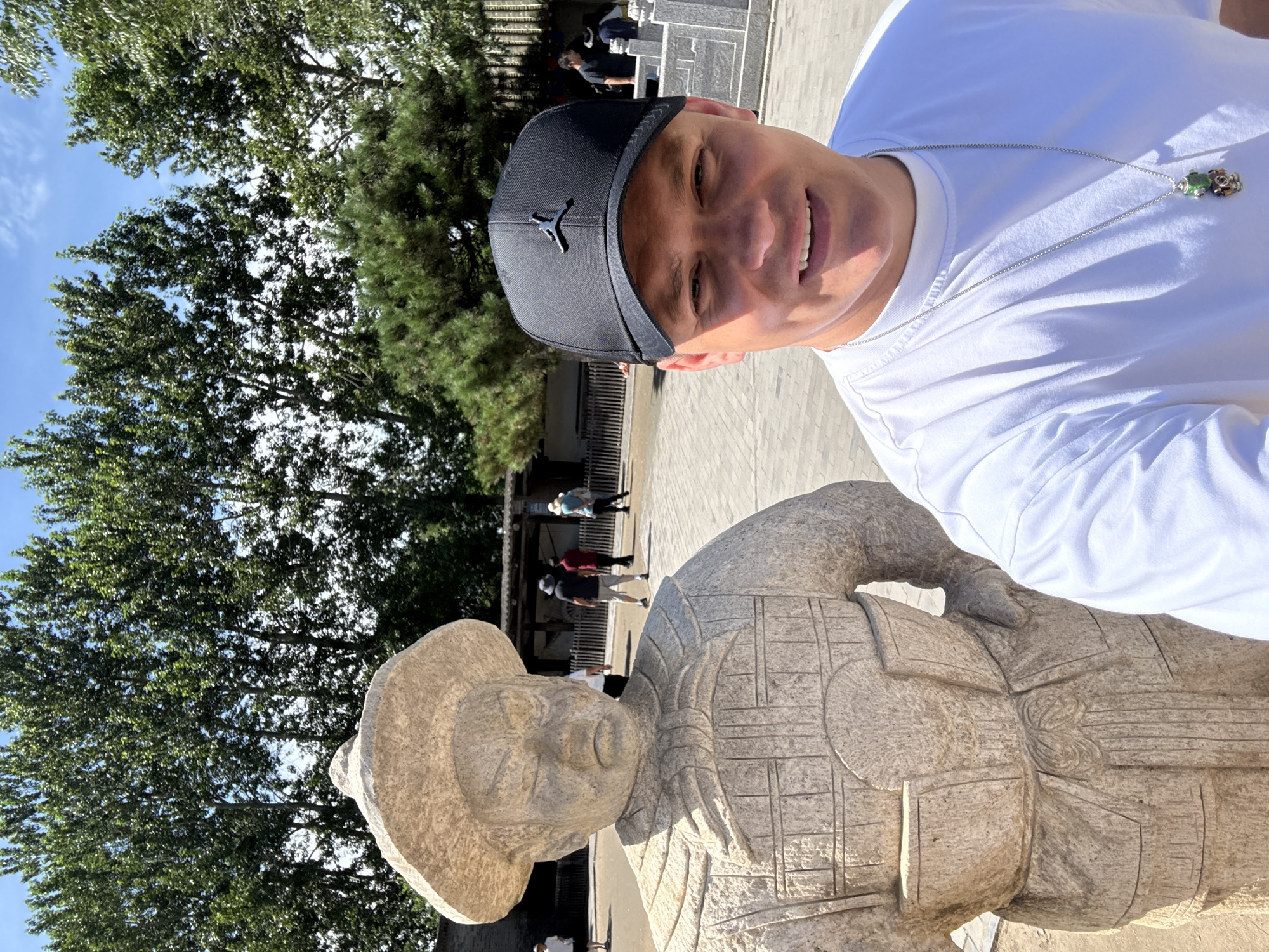 A man wearing a white t-shirt and a black baseball cap with a Jumpman logo is taking a selfie outdoors. To his left, a stone statue of a warrior in ancient armor and a hat stands prominently. In the background, other people are visible walking around a paved area with trees and traditional Chinese architecture. The scene appears to be in a historical site or park. Based on available information, this location is likely the tomb of Qin Shihuang, a significant historical site in Qinhuangdao, China.