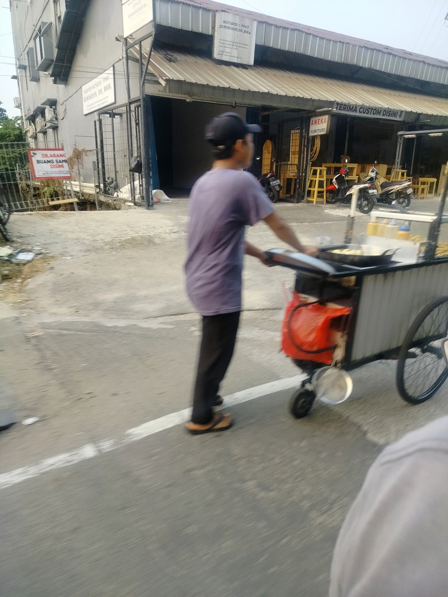 A low-angle, motion-blurred medium shot captures a street scene in East Jakarta, Indonesia, focusing on a man pushing a food cart. The man, wearing a purple t-shirt and dark pants, is seen from the side, his back partially to the camera, propelling a cart laden with food preparation equipment. The cart features a frying pan, bottles of condiments, and a red plastic bag. In the background, a building with several windows and air conditioning units is visible, along with a sign that reads "KANTOR NOTARIS PPAT SUNINGSIN, SH, MKN." Further in the background, a storefront with a sign saying "ANEKA FURNITURE" and "TERIMA CUSTOM DISINI" is present, along with several motorcycles parked outside. The ground is paved and shows a white line marking the edge of the road. The overall impression is that of a typical bustling street in a commercial area.