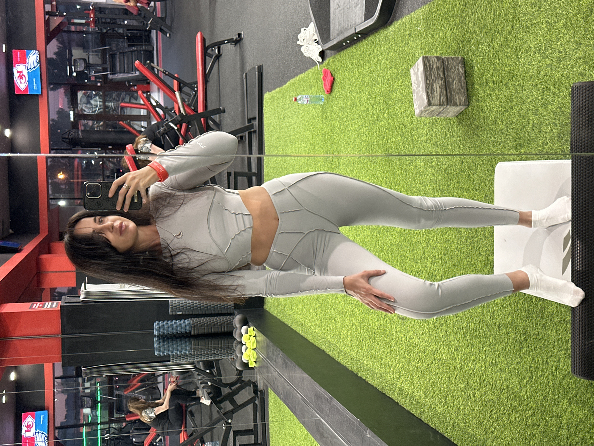 A woman is taking a mirror selfie in a gym in Irkutsk, Russia. She is wearing a light grey workout set consisting of a long-sleeved crop top and high-waisted leggings. The gym has a black floor with patches of artificial green turf. In the background, there are weight machines, benches, and other gym equipment. There are also mirrors and a television screen mounted on the wall. The overall lighting suggests it is either early morning or late evening.