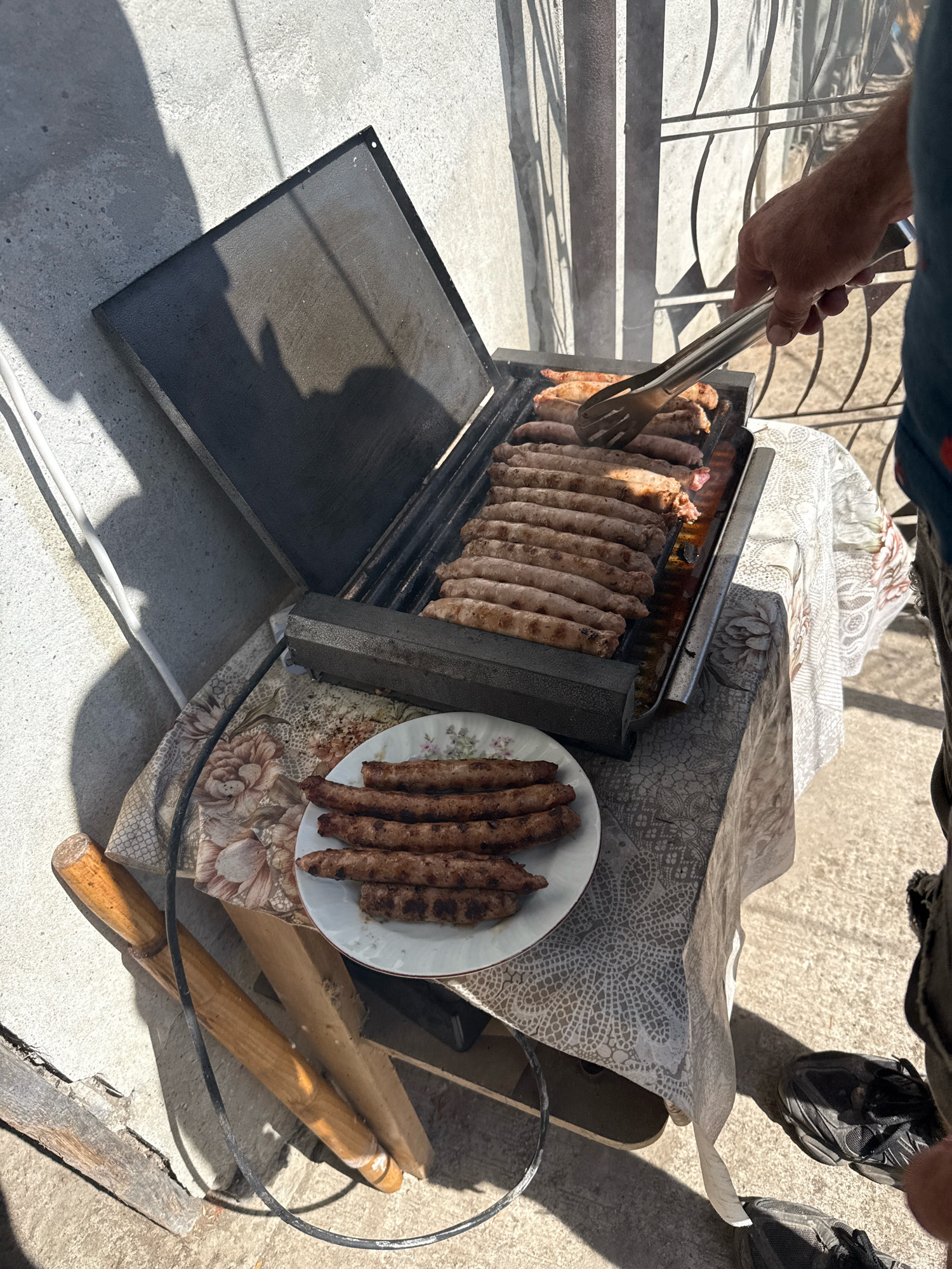In the city of Ekzarh Antimovo, Bulgaria, a close-up shot captures an outdoor grilling scene. The primary focus is a metal grill filled with numerous sausages, some of which are being handled by a person's hand using silver tongs. A plate of already grilled sausages sits to the right of the grill, placed on a table covered with a patterned tablecloth. The background reveals a concrete wall and a metal fence, illuminated by bright sunlight casting shadows on the ground. The overall setting suggests a casual outdoor meal preparation.