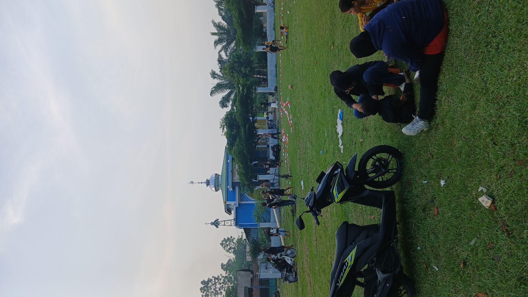 The image depicts an outdoor scene at Klumpang Kebun, Indonesia. In the foreground, two motorcycles are parked on a grassy field. A black scooter with lime green accents is positioned on the left, and another black motorcycle is partially visible on the right. Several people are seated and interacting in the middle ground. Three individuals, two adults and a child, are seated on the grass, facing away from the camera. One of the adults appears to be attending to the child. A person wearing a dark blue hoodie and red pants is also seated on the grass, facing towards the right. Further into the field, a group of individuals, some appearing to be children, are engaged in various activities, including what looks like a game of soccer with a visible orange ball. In the background, a blue mosque with a green roof and a silver dome stands prominently. Adjacent to the mosque are trees and a white fence. The sky is overcast, suggesting it might be late afternoon or early morning. Scattered red and white pieces of paper are visible on the grass in front of the mosque, possibly remnants of an event or decoration.