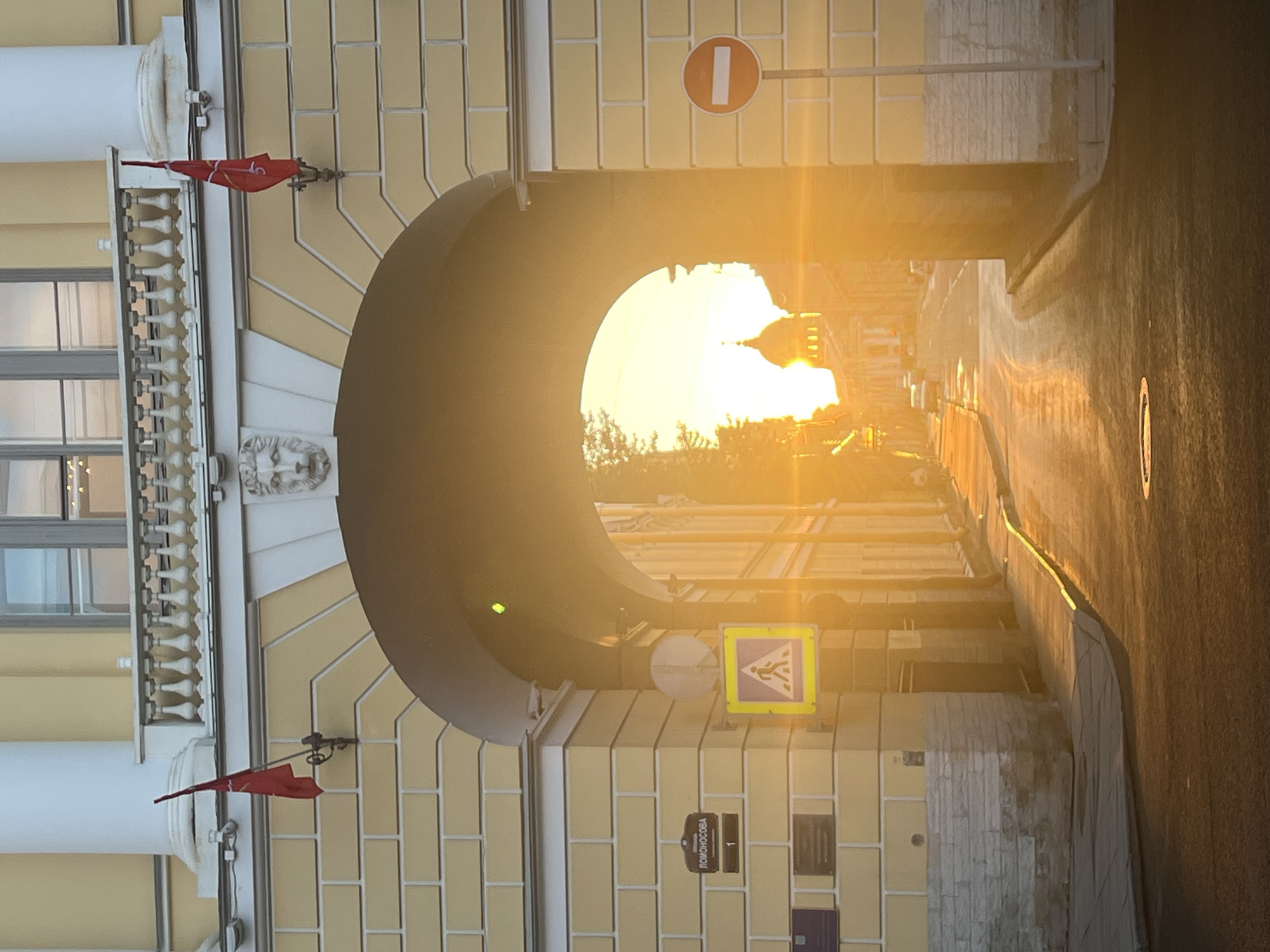 This is a vertical, outdoor photograph taken in Saint Petersburg, Russia. The sun is low in the sky, casting a warm, golden light that creates lens flare and silhouettes the scene. The light beams through a large arched passageway, illuminating a street lined with buildings. The archway is a prominent architectural feature, made of light-colored stone with a darker, curved interior.
On the left side of the archway, a light-yellow building with a white column and decorative railing is visible. Two small red flags are attached to the building, one near the top and one lower down. A decorative lion's head relief is visible on the wall. Windows with shutters are also present.
On the right side of the archway, the street curves away into the distance, reflecting the golden sunlight. A road sign with a red circle and a white horizontal bar, indicating "No Entry" in Russia, is mounted above the archway. Below the archway, a pedestrian crossing sign with a yellow background and a white silhouette of a person walking is mounted on a pole. A street sign with Cyrillic text reads "ПОНМОСООВА" and the number "1". Another sign, possibly a historical plaque, is also visible on the building wall.
The overall impression is one of a serene urban scene bathed in the beautiful light of either sunrise or sunset. The bright sunlight suggests clear weather. The architectural style and the road signs confirm the location as Russia.