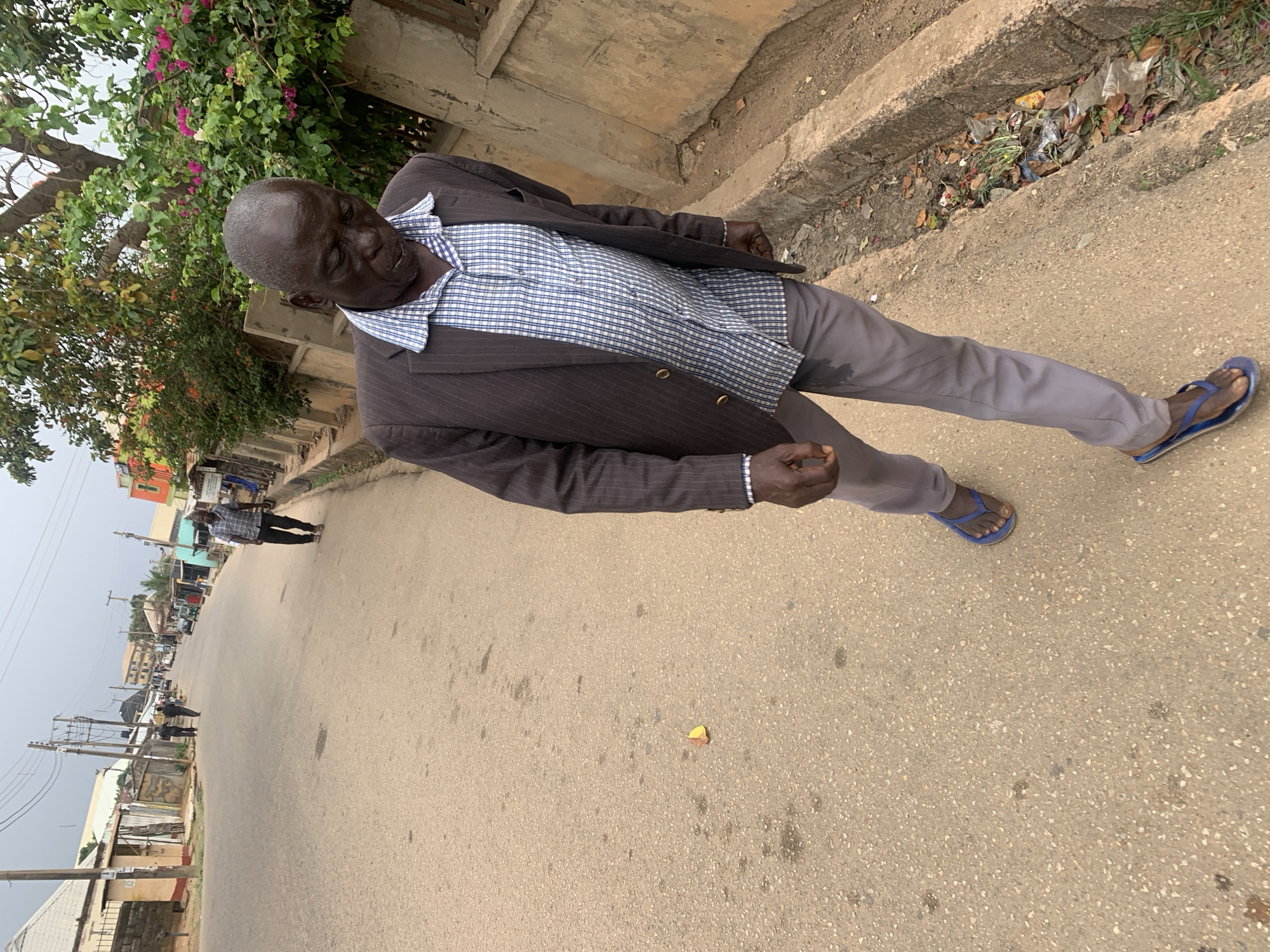 A man stands on a paved road in Jos, Nigeria, clad in grey slacks, blue flip-flops, a blue patterned shirt, and a brown suit jacket. He is positioned on the right side of the frame, facing the camera at an angle. In the background, a streetscape extends into the distance, with buildings and utility poles visible. A concrete curb with some litter is visible on the right. A tree with green foliage and pink flowers is present in the upper left corner. Another person is walking in the distance to the left of the primary subject.