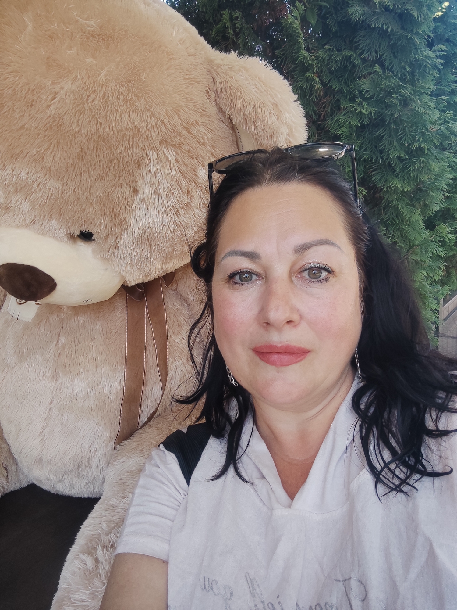 A middle-aged woman with dark, wavy hair and light skin poses for a selfie next to a large, light brown teddy bear. She is wearing a light-colored, possibly white, t-shirt and has sunglasses perched on top of her head. The teddy bear is positioned behind her, filling a significant portion of the frame. In the background, lush green foliage, likely from a tree, is visible. The scene appears to be outdoors, possibly in a park or garden setting within Sochi, Russia. The lighting suggests it is daytime.