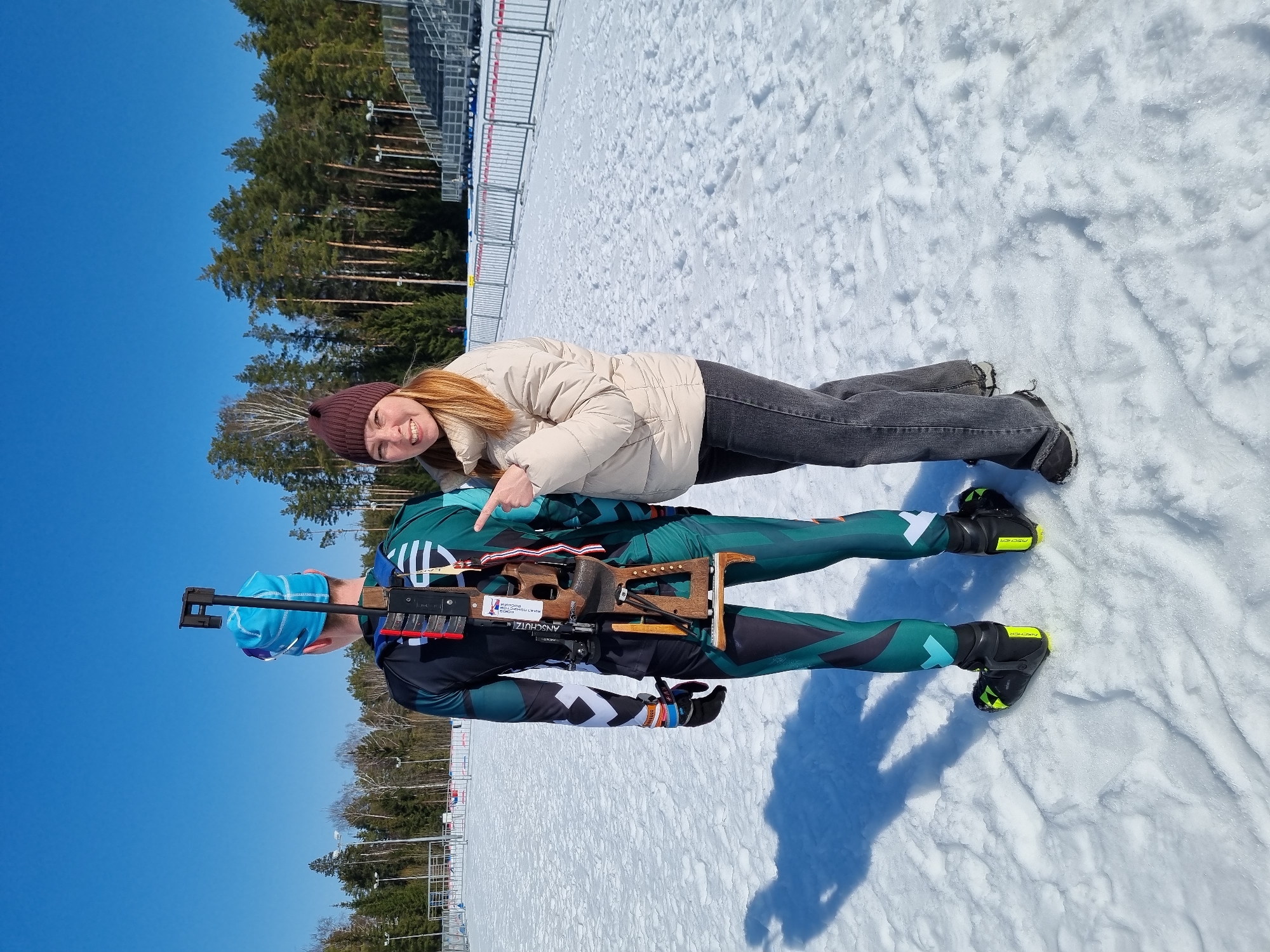The image captures an outdoor winter scene in Прикамский, Russia, at what appears to be a biathlon or cross-country ski complex under a clear blue sky. A male individual, identified as a biathlete, stands with his back largely towards the camera, dressed in a form-fitting dark teal, black, and white biathlon suit, a light blue beanie, and black ski boots with neon yellow trim (Fischer branded). A biathlon rifle (Anschutz branded, with a wooden stock and black components) is strapped vertically to his back. To his immediate right, a female individual stands, partially facing the camera with a smile. She wears a beige puffy winter jacket, a dark brown knit beanie, dark grey jeans, and dark winter boots. Her right arm is extended, with her index finger pointing towards the biathlon rifle on the male's back. The ground is covered in white snow with visible footprints. In the background, a dense line of tall pine trees is visible, along with grey spectator bleachers and red and white temporary safety barriers lining a snow track. The scene is illuminated by bright sunlight, casting shadows on the snow.