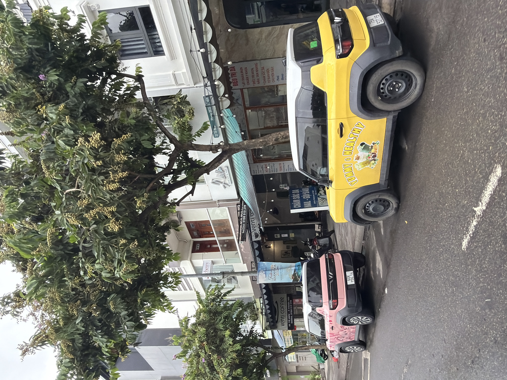 The image depicts a street scene in Nha Trang, Vietnam. Two small, modern cars are parked on the right side of a wet, asphalt road. The car in the foreground is bright yellow with a white roof and a decal on its side featuring cartoon images of fruits and vegetables with the text "YUMMY & HEALTHY" in yellow lettering. Its license plate is visible and reads "79A 527.19". The car behind it is pink with white accents and also has decals on its side, featuring cartoon characters. Its license plate reads "60K 156.14".
In the background, there are several buildings lining the street. Some of these buildings appear to be shops or businesses, indicated by awnings and signage. One sign reads "VENTI COFFEE". Trees with green leaves and small, light-colored fruits or blossoms are prominent, casting shadows on the buildings and street. The sky is overcast. The overall impression is a quiet street scene in an urban environment.