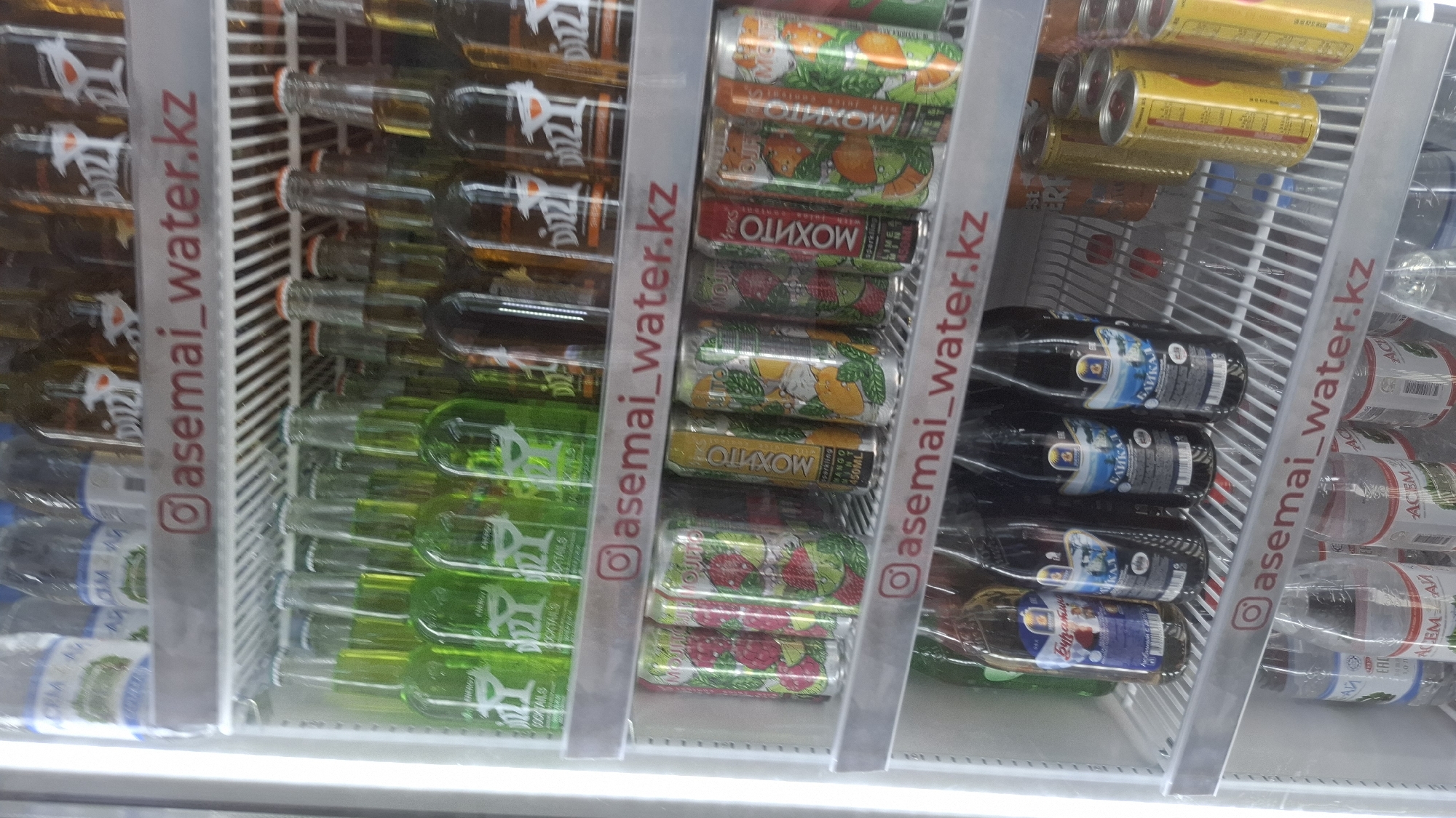 The image displays the interior of a refrigerated display case stocked with various beverages. The case contains multiple shelves filled with bottles and cans of drinks. On the top shelf, there are several green bottles with white labels featuring a stylized glass graphic, possibly representing cocktails. Next to these are brown bottles. Below this shelf, the Instagram handle "@asemai_water.kz" is visible, printed in red on a white background, indicating this is likely a business in Shymkent, Kazakhstan. The next shelf down holds an assortment of aluminum cans. Some cans have colorful designs featuring fruit like raspberries and limes, and text that appears to be "MOJITO". There are also yellow cans with text. Further down, more bottles are visible, including some dark colored ones with Cyrillic labeling that appears to say "BAIKAL" and another bottle with Cyrillic text that says "Бураны". The bottom shelf shows clear plastic bottles with blue and white labeling. The overall environment is clean and organized, typical of a retail beverage display.