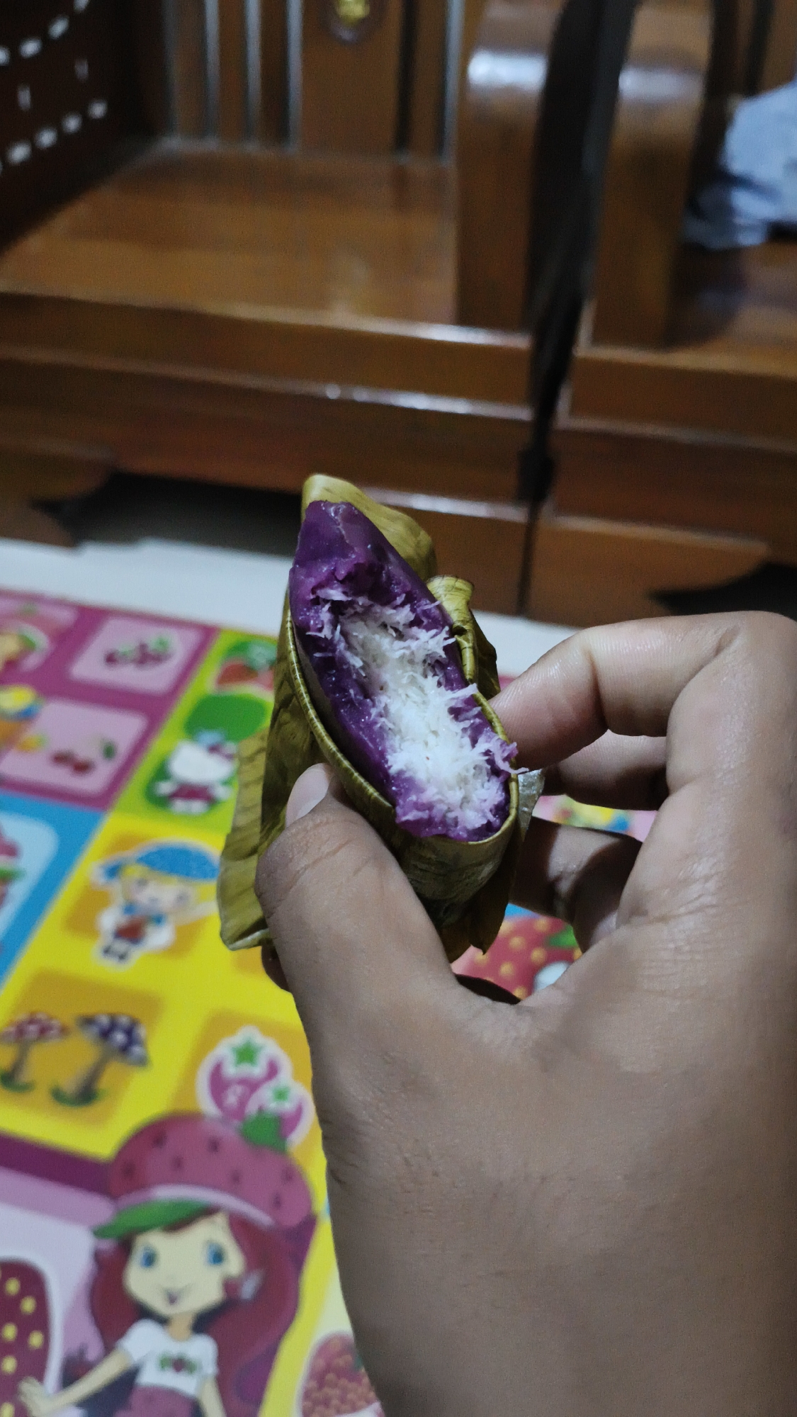 A person holds a Filipino delicacy, likely a Puto Bumbong, made with purple glutinous rice and filled with shredded coconut. The food item is wrapped in a banana leaf. The background consists of a wooden chair and a colorful mat with cartoon characters. The scene appears to be indoors.