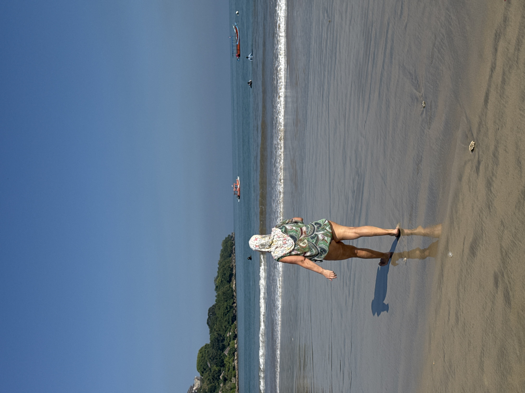 A sunny, horizontal, medium-range shot shows a woman walking on a beach in Kuta Selatan, Indonesia. The woman, facing away from the viewer and to the right, wears a green and white patterned outfit and a sunhat. She casts a shadow on the wet sand. The beach curves gently, with waves breaking on the shoreline. In the background, the blue sea merges with a clear blue sky. Several small boats and what appears to be at least one person swimming are visible in the water. Along the left edge of the frame, a dense, green forest covers the land, indicating a coastline.