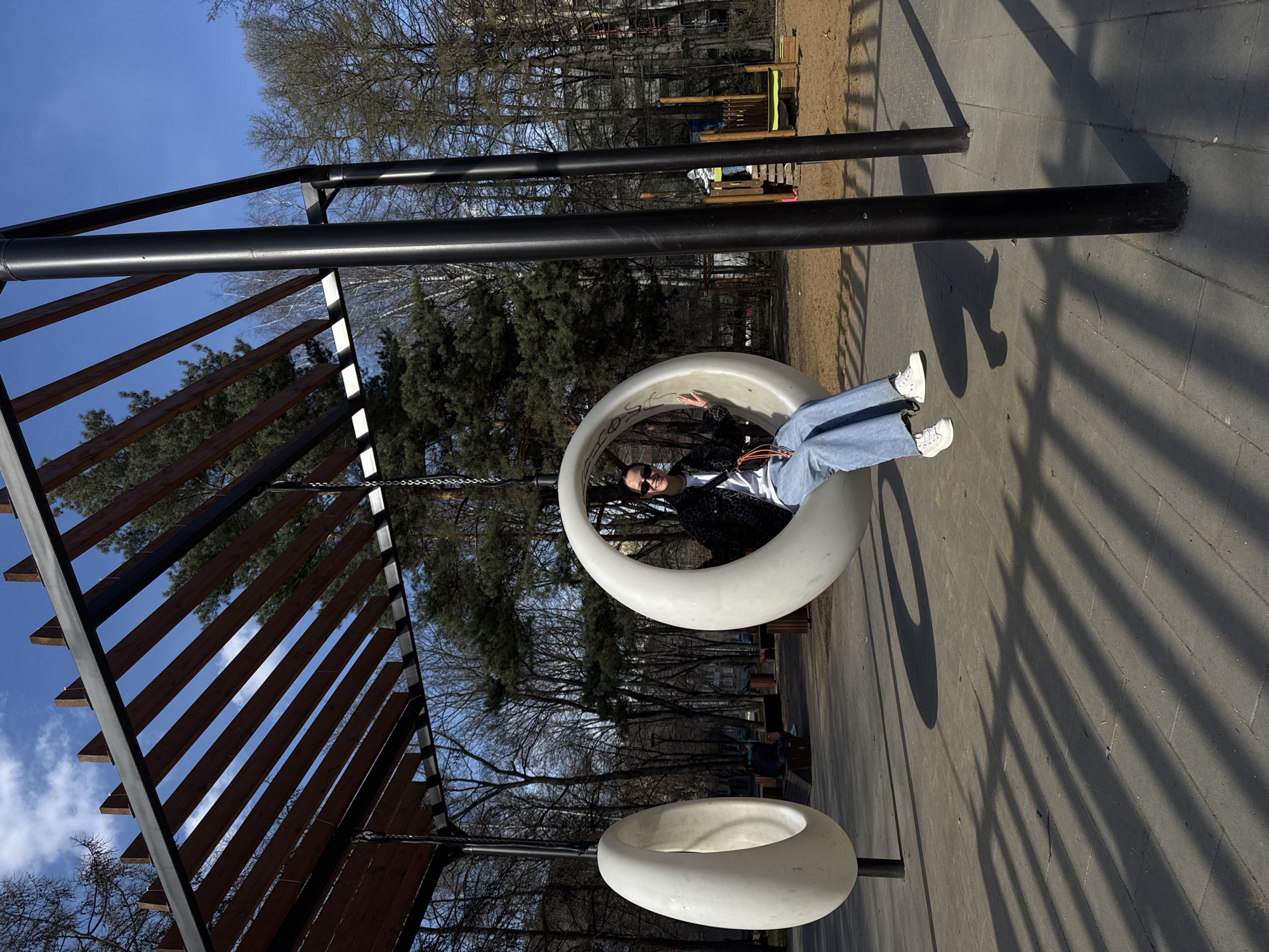 A park scene, seemingly in Moscow, Russia, presents a woman seated in a suspended, circular swing. The playground features a black metal frame supporting the swing and wooden slats forming a partial canopy. The ground is paved with concrete tiles casting striped shadows. Trees with bare branches frame the scene, and hints of other playground structures are visible in the background. The woman wears jeans, a white shirt, and a black jacket, with sunglasses obscuring her eyes.