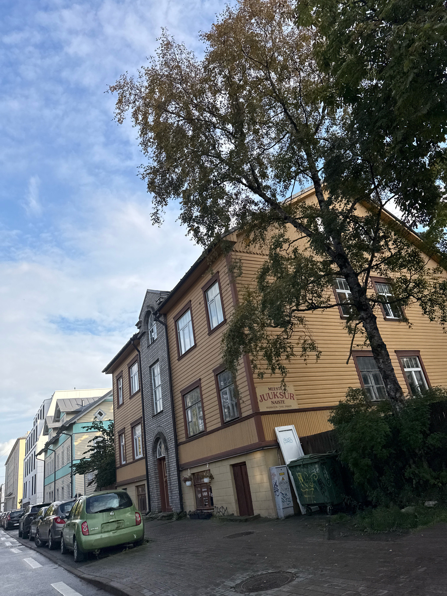 The image depicts a street scene in Tallinn, Estonia, with a focus on several buildings and parked cars. In the foreground, a bright green hatchback, identified as a Nissan Micra, is parked curbside. Adjacent to it are other cars, mostly sedans and hatchbacks, lined up along the street. The buildings lining the street exhibit a mix of architectural styles. A prominent yellow wooden building with horizontal siding stands to the right, featuring multiple windows and a sign with Estonian text, "MEESTE JUUKSUR NAISTE MANIKUUR NAIL DISAIN." To its left is a narrower gray stone building with a mansard roof and arched window details. Further down the street, other buildings with varying colors and styles are visible, including a modern white building with horizontal elements and a pastel blue and white structure with decorative gables. A large tree with sparse autumn foliage leans over the yellow building, casting shadows. Two large green dumpsters and a white upright panel are situated near the base of the yellow building, showing signs of graffiti. The sky is a mix of blue with scattered white clouds, indicating daytime. The street appears to be paved with cobblestones, and a manhole cover is visible in the lower left corner of the frame. The overall impression is of a residential or mixed-use urban environment in Tallinn.