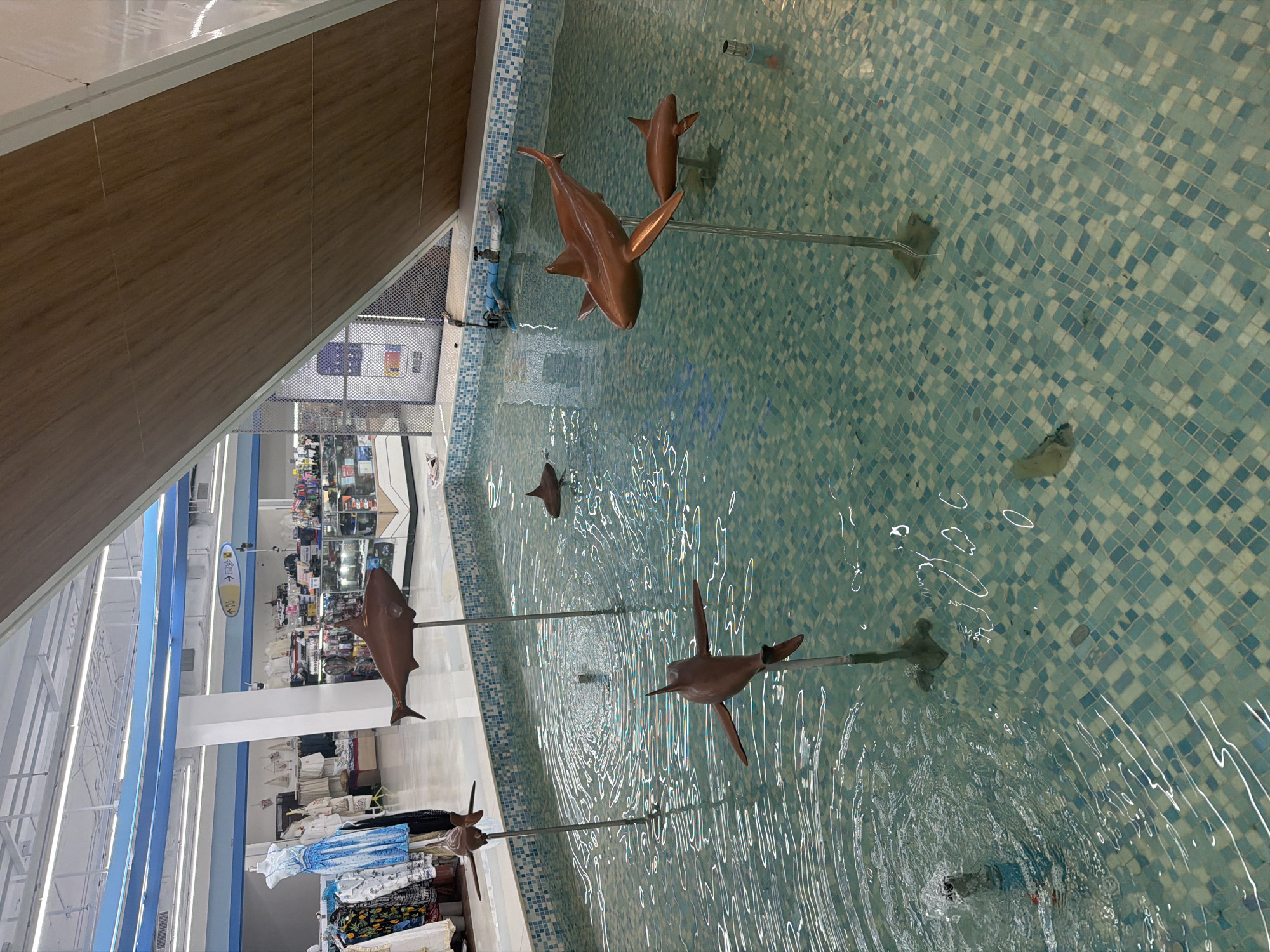 The image depicts an indoor water feature at a retail location. The water basin is lined with blue and white mosaic tiles and contains several bronze-colored shark statues mounted on vertical poles. The water is rippled, suggesting active circulation.
In the background, through an opening, there's a view into the retail space, including store lighting, shelving, and merchandise displays. Some clothing items are seen in the foreground.