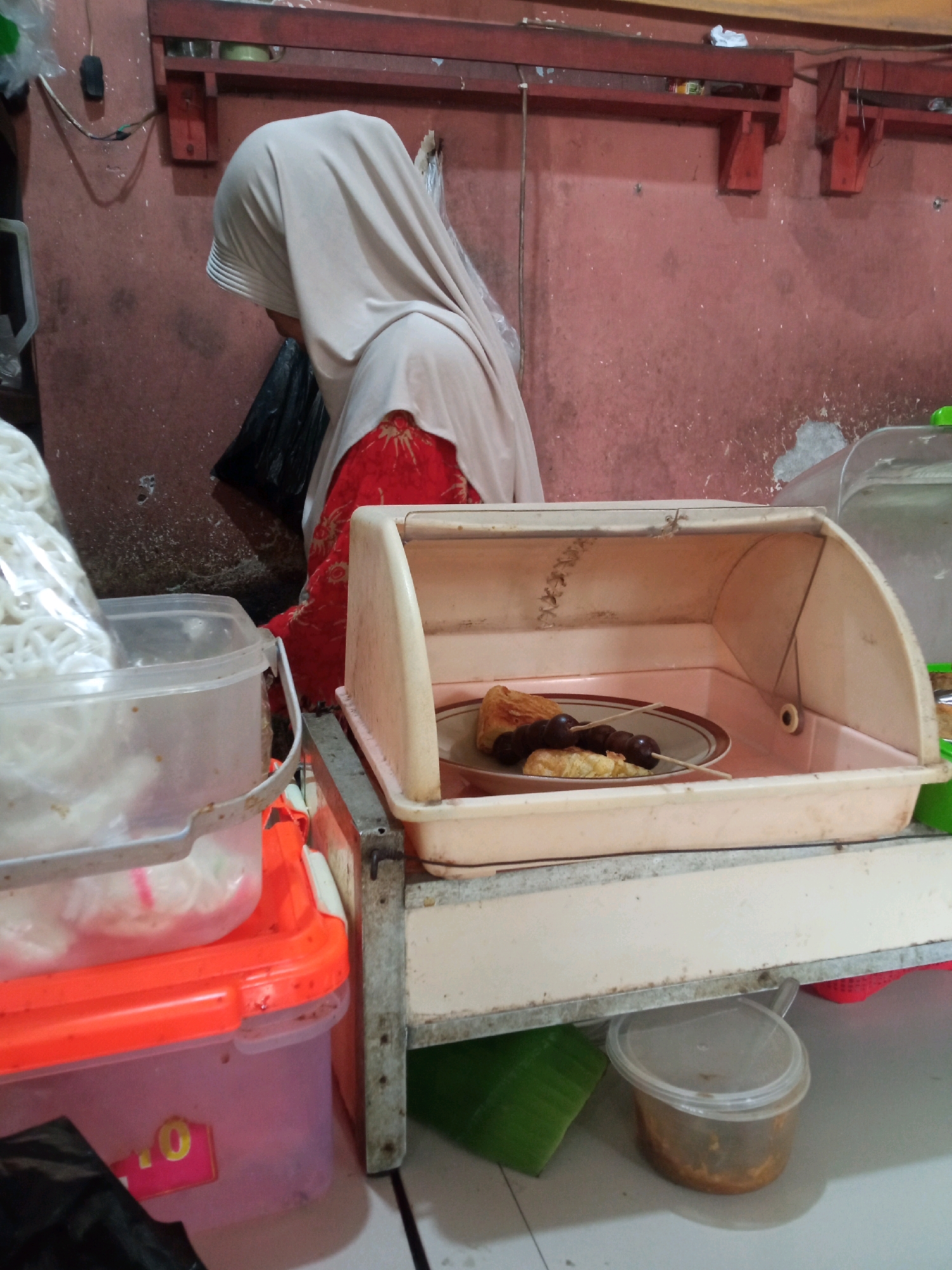 An indoor, eye-level, medium shot captures a stall scene at night, likely in Sambong, Indonesia. A woman wearing a light-colored hijab and a red patterned garment is partially visible from the right side of the frame, facing away from the camera. Her posture suggests she is attending to her stall.
In the foreground, a light-colored plastic display case sits on a metal stand. Inside the case, a plate holds two pieces of pastry, one triangular and one square, skewered with two dark meatballs. The display case is open, revealing the food within.
To the left of the display case, a clear plastic container holds a pile of white, noodle-like food. Below this, an orange plastic container is stacked on top of a pink plastic container. A black plastic bag is visible at the bottom left of the frame.
The background features a reddish-pink wall and a wooden shelf unit. The environment appears to be a food stall or market setting. The lighting suggests an artificial light source illuminating the scene.