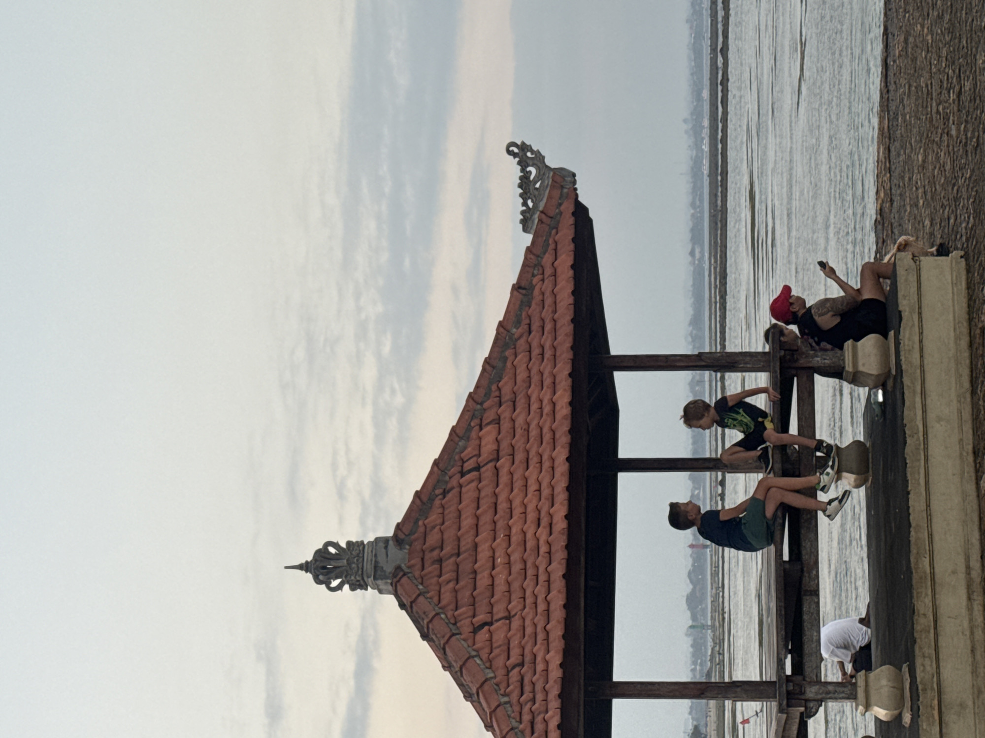 The image depicts a pavilion with a tiled roof and ornate carvings on its eaves. Inside the pavilion, two young boys are seated on a wooden platform, facing each other and engaged in conversation. To the left, another individual wearing a white shirt is crouched down. To the right, a person in a red cap is seated, holding a mobile phone. The pavilion is situated beside a body of water, with the shoreline visible in the background. The sky is overcast with clouds. This scene is likely located in Denpasar, Indonesia, given the architectural style of the pavilion, which is characteristic of Balinese design.
