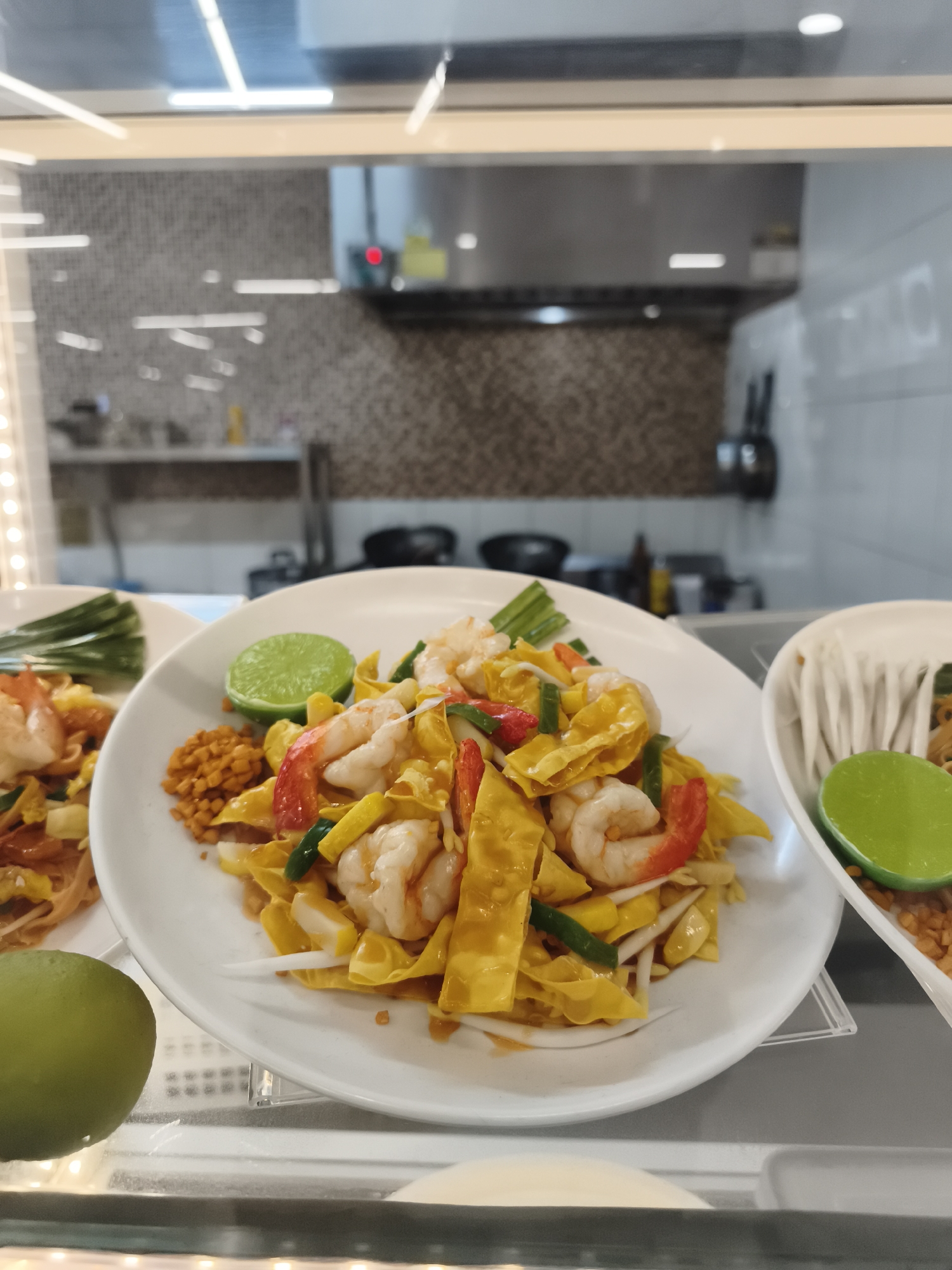 The image shows three plates of Pad Thai, a stir-fried rice noodle dish, displayed under a glass counter. The dishes include shrimp, tofu, sprouts, lime wedges, and other ingredients commonly found in Pad Thai. The backdrop suggests a kitchen or food preparation area with tiled walls and stainless steel equipment. It is unclear if this image was taken in Baan Lamai, Thailand as it would need additional information to make that determination.
