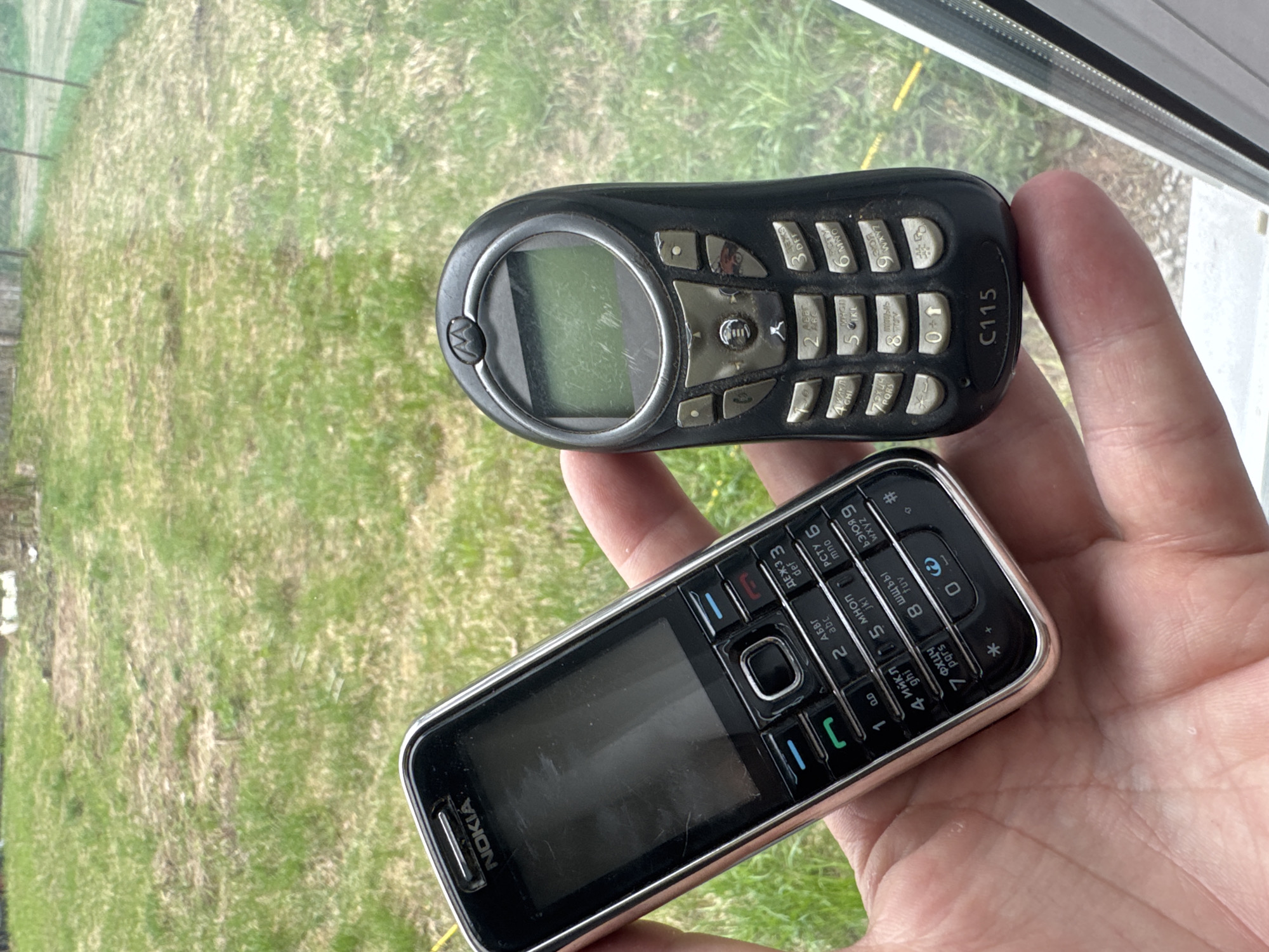 A person's hand holds two old cellular phones, a Motorola C115 and a Nokia phone. The Motorola is on top and is black with a round screen and push-button keypad. The Nokia phone is silver-trimmed and black with a rectangular screen and push-button keypad. The background is a blurry view of a grassy area, possibly seen through a window. The location is Chuprino, Russia.