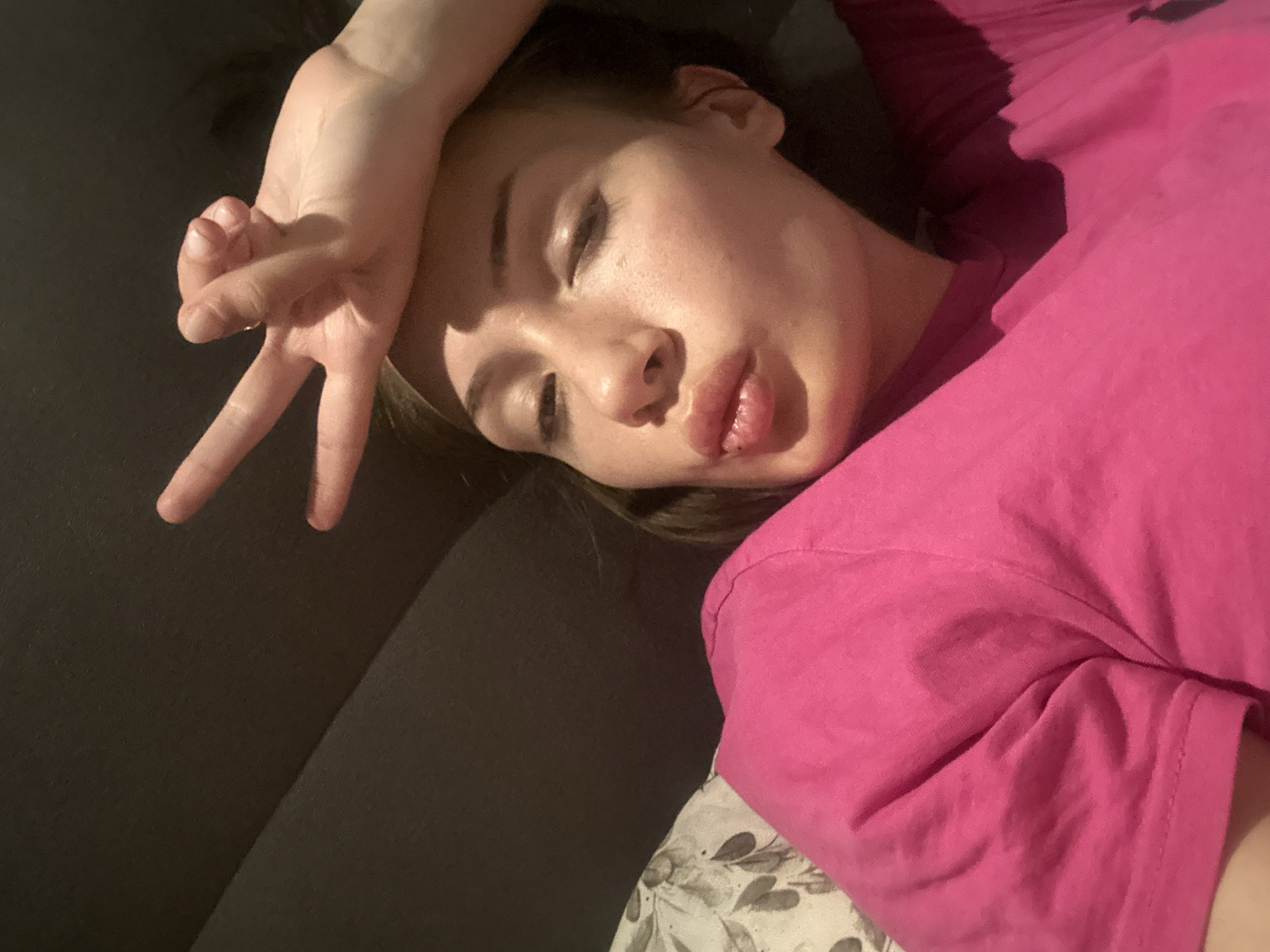 A woman is lying down, her head resting on a dark surface, likely a couch or bed. She is wearing a bright pink t-shirt. Her face is angled towards the camera, with her eyes partially closed. One hand is raised, fingers forming a V-shape. The lighting is uneven, casting shadows across her face and the surrounding area. The bedsheets are underneath the woman and contain a white floral design. It is impossible to determine if the picture was taken in Burgas, Bulgaria or any other city based solely on the provided image.