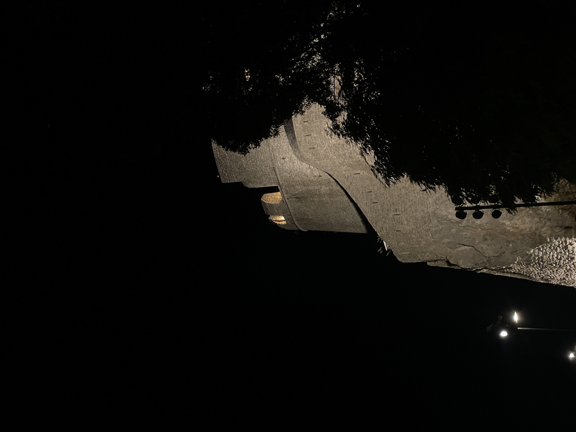 The image shows a nighttime view of the Kanli Kula Fortress in Herceg Novi, Montenegro. The fortress, constructed from stone, is illuminated by spotlights, highlighting its textured walls and ramparts. A prominent round tower with arched openings is visible. The dark sky provides a stark contrast to the lit structure. In the foreground and to the right, the silhouette of a large tree partially obscures the fortress. On the left side of the image, streetlights are visible at the bottom, indicating a nearby urban setting. The scene is still and quiet, with no people or animals present. The overall impression is one of historical architecture set against a dark, nocturnal landscape.