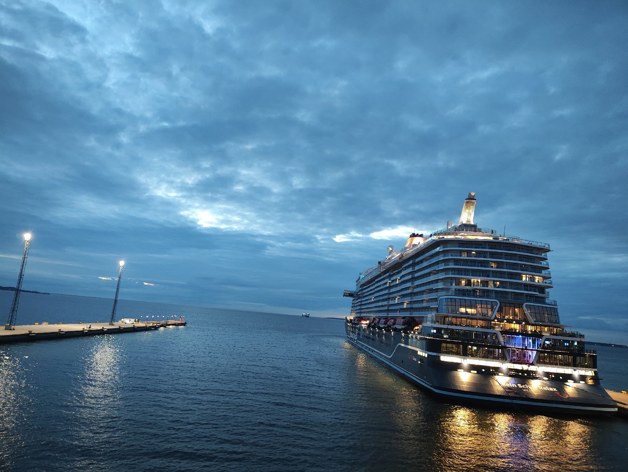 An uncropped, eye-level, wide shot captures a large cruise ship, the "Mein Schiff 1", docked in Tallinn, Estonia, at dusk. The ship, seen from its stern, is illuminated by numerous lights, reflecting off the calm, dark water. The sky is filled with dramatic, moody clouds, with patches of lighter sky visible. In the foreground, the dark blue water has rippling reflections of the ship's lights. To the left, a pier stretches out into the water, equipped with two tall light poles that cast bright white beams onto the pier and the water. The pier has several bollards and a small building or kiosk. In the distance, the outline of land is faintly visible. Two smaller ships are also visible on the horizon. No people are clearly discernible in the image. The overall scene conveys a sense of calm and stillness at the end of the day.