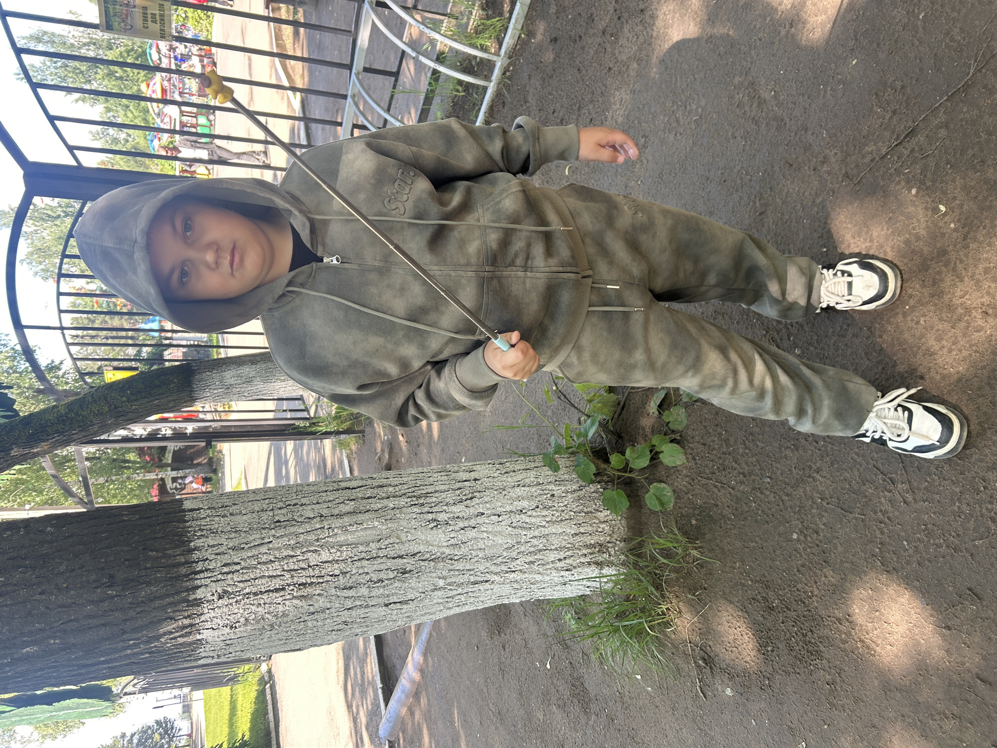 An outdoor, eye-level full shot captures a young child standing on a dirt path in what appears to be a park or playground in Chaikovsky, Russia. The child, with a neutral expression and light-colored eyes, looks directly at the camera. They are dressed in a hooded, olive green or khaki full-zip tracksuit, which has a distressed or tie-dye effect. The word "Star" is visibly embroidered on the left chest area of the hoodie. The child's hood is up, framing their face. They are wearing white and black chunky sneakers. In their right hand, they hold a thin, light blue-handled stick or fishing rod-like object with a yellow tip.
Behind the child, a black metal fence or railing separates the immediate area from a background showing green trees and colorful amusement park rides or playground equipment. There's a clear blue sky overhead, and bright sunlight casts shadows on the ground, indicating it's daytime. To the left of the child, two large tree trunks stand, one lighter in color than the other. Grass and small plants grow near the base of the trees and along the path. In the background, beyond the fence, a yellow sign with Cyrillic text is visible, which reads "СТОЯНКА ДЛЯ ВЕЛОСИПЕДОВ" (BICYCLE PARKING).