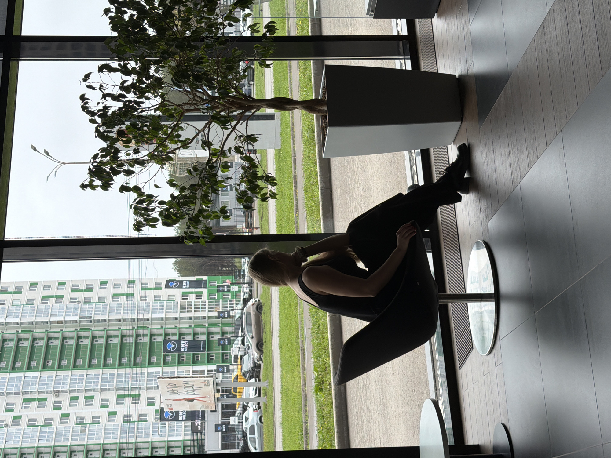 The photograph depicts a woman sitting in a dark-colored chair next to a potted tree. The setting appears to be an interior space, possibly a cafe or a waiting area, with a large window offering a view of the outside.
Outside the window, there is a multi-story building with green and white walls, typical of residential buildings. There are several parked cars and greenery suggesting a street or parking area.
Based on the signage visible through the window, the location can be pinpointed to Izhevsk, Russia.