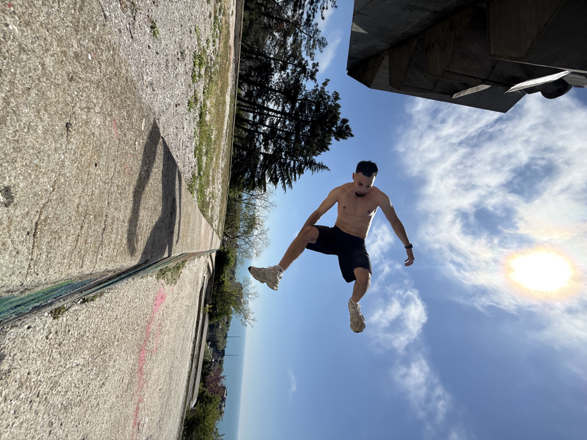 A shirtless man with dark hair and shorts is jumping in the air with a blue sky and clouds as a backdrop. He is wearing running shoes. The camera angle is tilted. Concrete architecture and trees are visible in the background. A body of water is visible. A portion of the Monument to the Bulgarian-Soviet Friendship in Varna, Bulgaria is in the background.