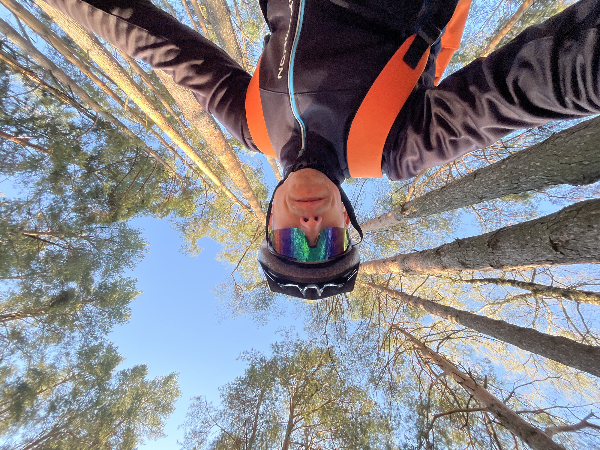 A low-angle, upward-facing photograph shows a person in cycling gear in a forest. The person is wearing a black cycling jacket with orange shoulder straps and a blue stripe down the front. The person also wears a helmet and reflective sunglasses. The background is dominated by tall trees that stretch upwards, with foliage and branches interspersed against a blue sky. The scene appears to be bathed in sunlight. The person is in the city of Chaikovsky, Russia.