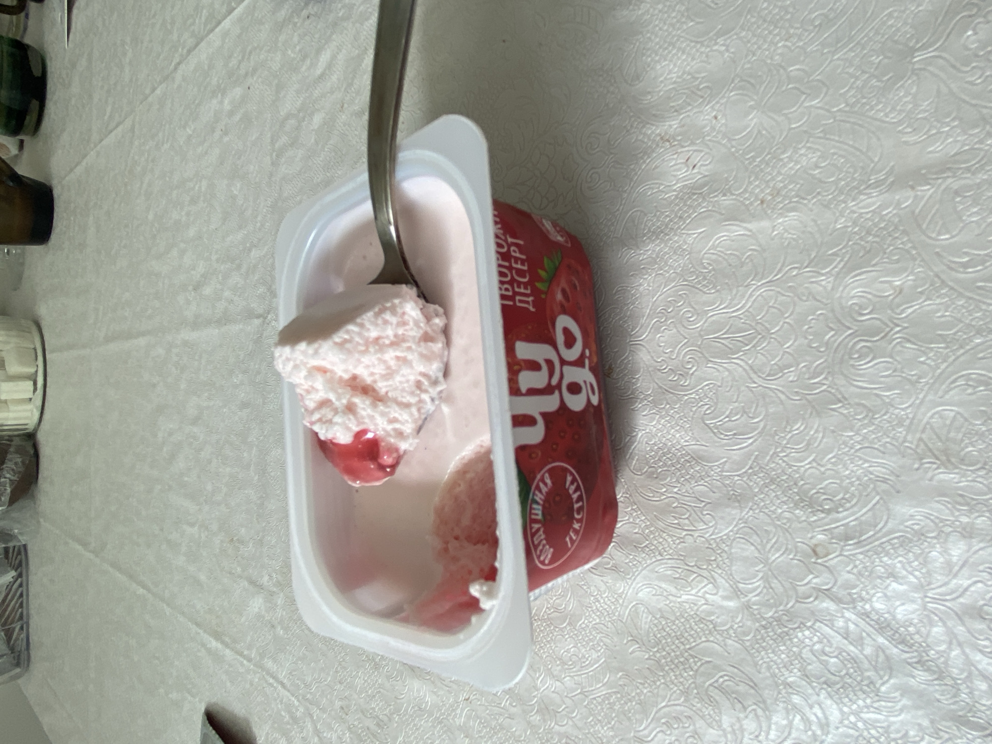 The image, taken in Chaikovsky, Russia, features a close-up of a white plastic container holding a pink, whipped, strawberry-flavored dairy dessert. A silver spoon is prominent, lifting a generous scoop of the dessert which also includes a small amount of darker red, jam-like fruit filling. The dessert container has a vibrant red label on its side, displaying the brand name "Чудо" (Chudo, meaning Wonder) and the text "ТВОРОЖНЫЙ ДЕСЕРТ" (Tvorozhnyy Desert, meaning Curd/Quark Dessert). A circular graphic on the label further specifies "ВОЗДУШНАЯ ТЕКСТУРА" (Vozdushnaya Tekstura, meaning Airy Texture) and "КЛУБНИКА" (Klubnika, meaning Strawberry). The container is placed on a white tablecloth featuring an intricate, embossed or woven floral and damask pattern. In the blurry background to the left, glimpses of dark-colored glasses or bottles are visible. The scene is set indoors, with bright lighting indicative of daytime.