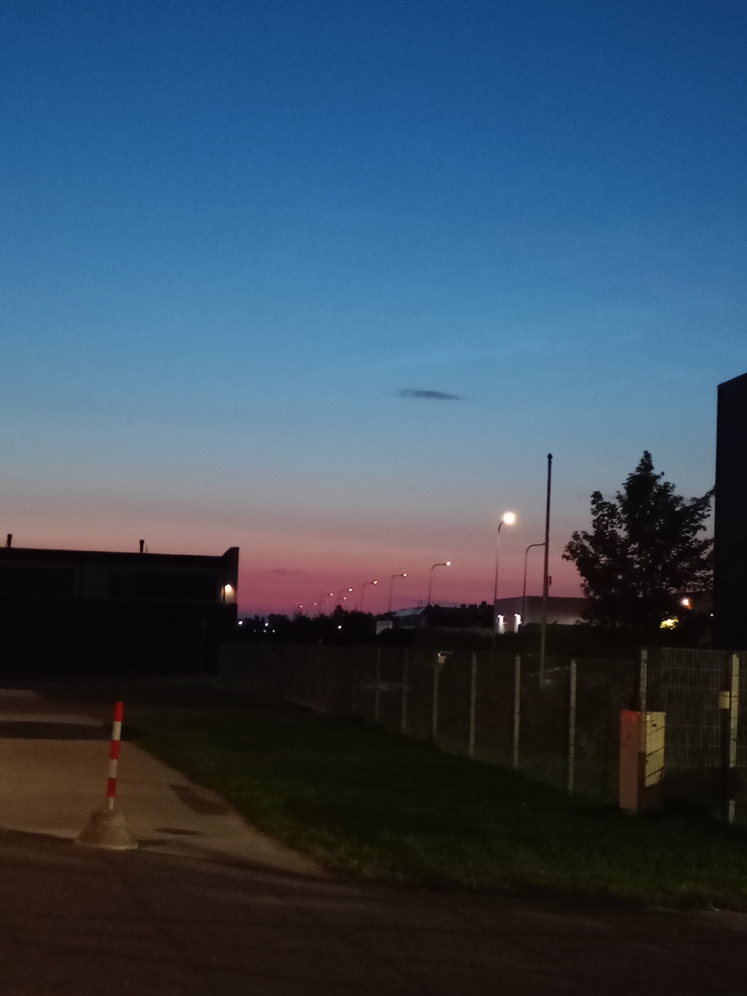 The image depicts an exterior scene at dusk in Peetri alevik, Estonia. The sky transitions from a deep blue at the top to shades of pink and orange near the horizon. In the foreground, a traffic cone with red and white stripes is positioned on a paved surface. To the left, a dark building with illuminated windows is visible. A fence with vertical posts runs horizontally across the mid-ground, separating a grassy area from the foreground. Several streetlights are present, some illuminated, along with a single tree with dark foliage. In the background, more buildings and lights suggest an urban or suburban environment.
