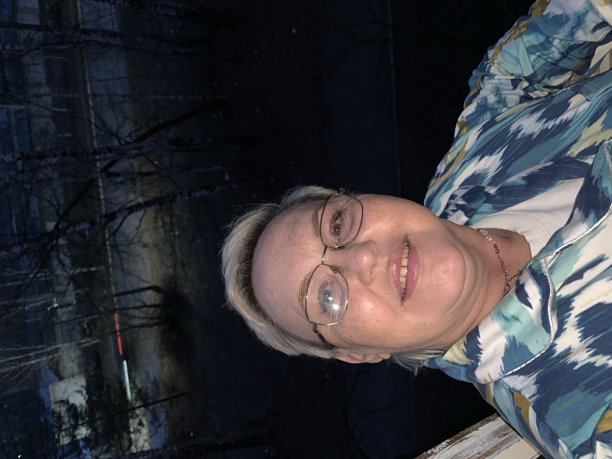 An image, rotated 90 degrees counter-clockwise, depicts a middle-aged woman posing for a photo outdoors at night in Chaikovsky, Russia. The woman, with light blonde or grey hair, is wearing gold-rimmed glasses that show bright reflections from a light source, likely a camera flash. She has visible freckles and is smiling faintly, looking directly at the camera. She is dressed in a vibrant, patterned jacket featuring shades of blue, white, and a muted gold or light brown, worn over a white ribbed top. A delicate chain necklace is visible around her neck.
The background is very dark, indicating nighttime. Silhouettes of bare tree branches are prominent against the dimly lit distance. Through these branches, the faint outline of a building can be discerned, characterized by vertical streaks of red and white light, suggesting artificial illumination from the structure or nearby sources. A white railing is visible in the lower right foreground, suggesting the woman might be on a balcony or porch. No visible text is present in the image.